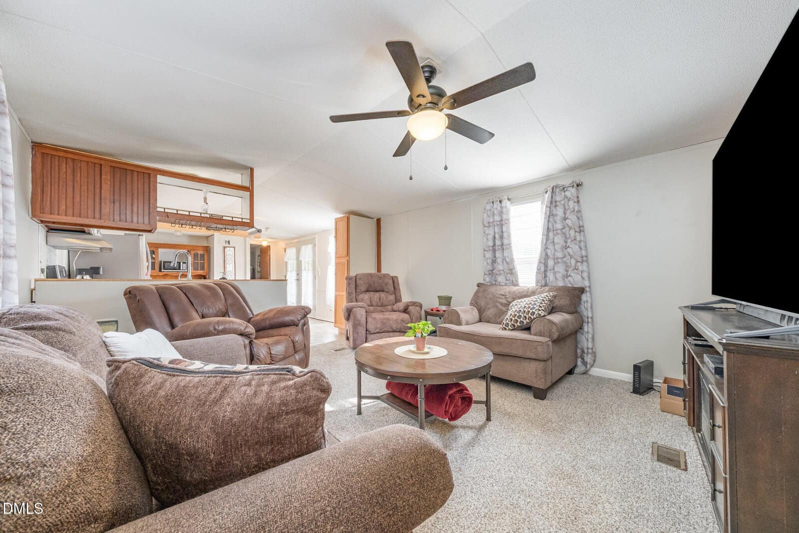 A living room with brown upholstered furniture, a round coffee table, a large flat-screen TV, a ceiling fan with lights, and neutral walls with a window covered by patterned curtains. The kitchen is visible in the background.