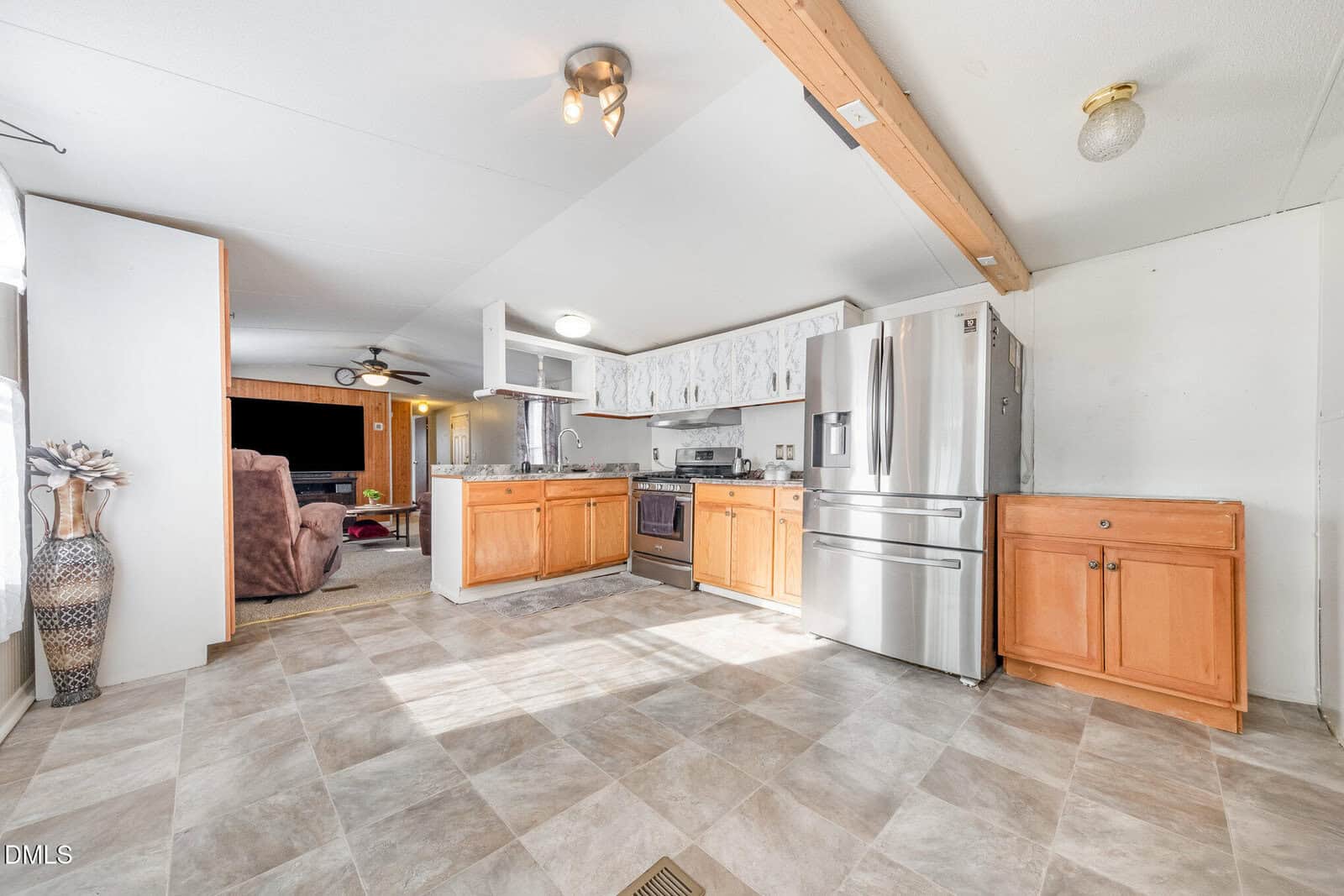 A spacious kitchen with light-colored tile flooring, stainless steel appliances, wood cabinets, marble-patterned upper cabinets, and a wooden ceiling beam. An adjoining living area with a brown recliner is visible.