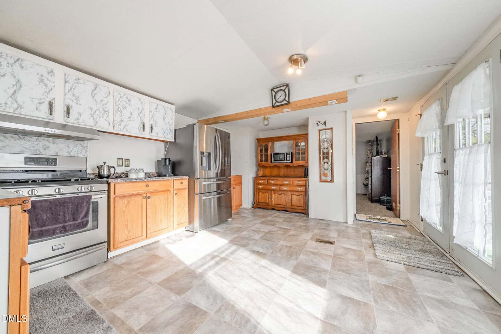 Bright kitchen with stainless steel appliances, wood and white cabinets, tiled floor, a wooden buffet, and double glass doors with sheer curtains letting in natural light. A hallway is visible in the background.