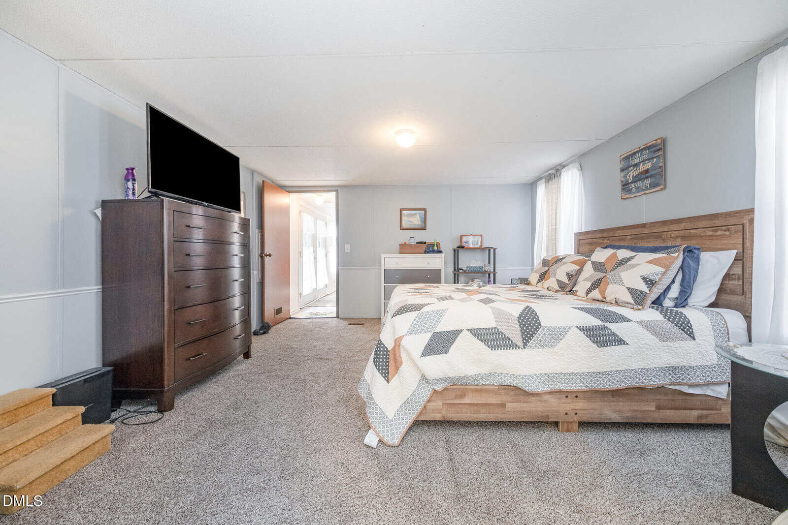 A bedroom with a wooden bed, a geometric-patterned quilt, a dark wood dresser with a TV on top, a desk with items, carpeted floor, and light gray walls. Natural light enters through covered windows.