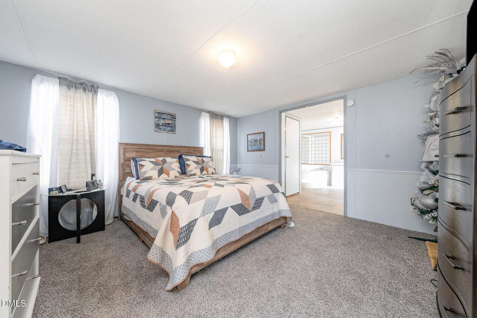 A bedroom with light blue walls, a queen-sized bed with a geometric-patterned quilt, two windows with sheer curtains, a dresser, a nightstand, and an open door leading to an adjacent room.