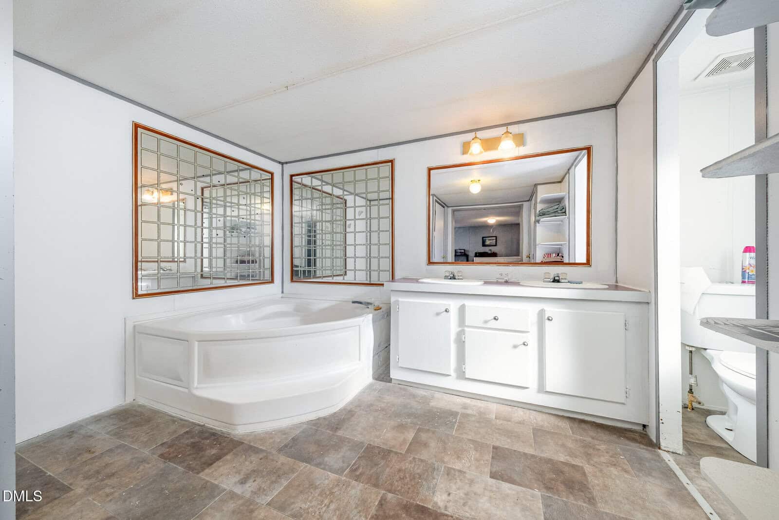 Spacious bathroom with a corner bathtub, two large mirrors, white double-sink vanity, and shelves. Toilet is visible in a nook to the right. The floor has a brown tile pattern.