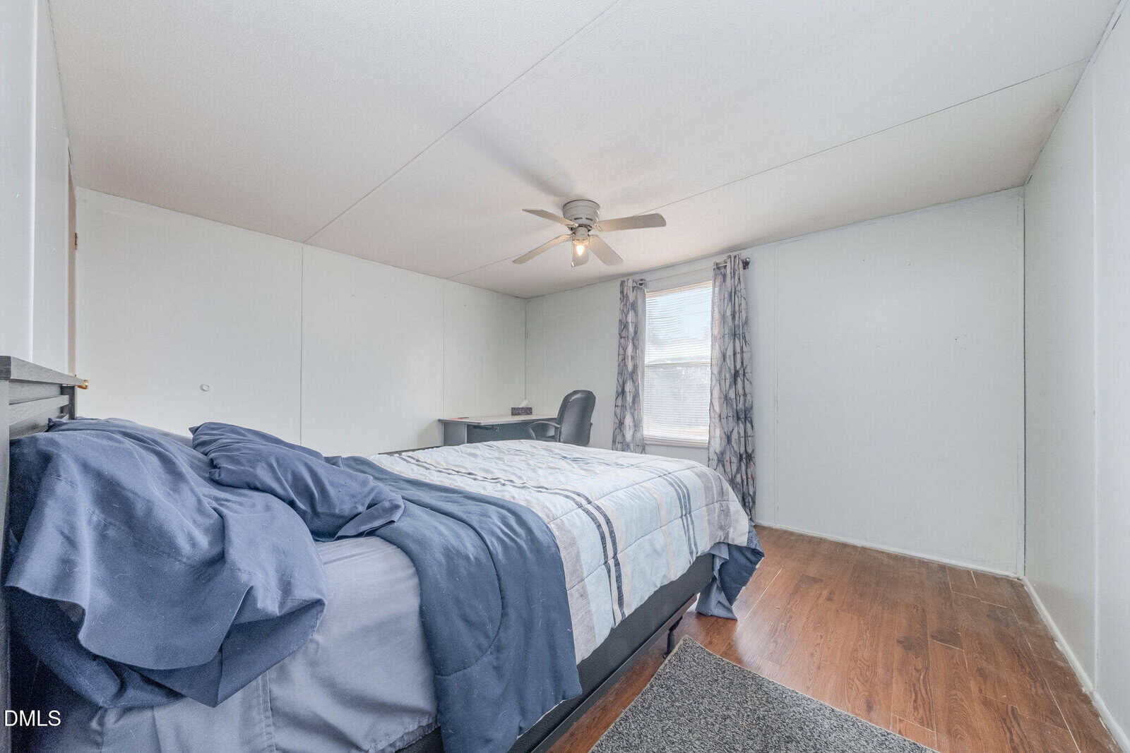 A simple bedroom with a bed covered in blue and gray bedding, a desk with a chair by the window, two patterned curtains, a ceiling fan, and hardwood floors. The walls are plain and light-colored.