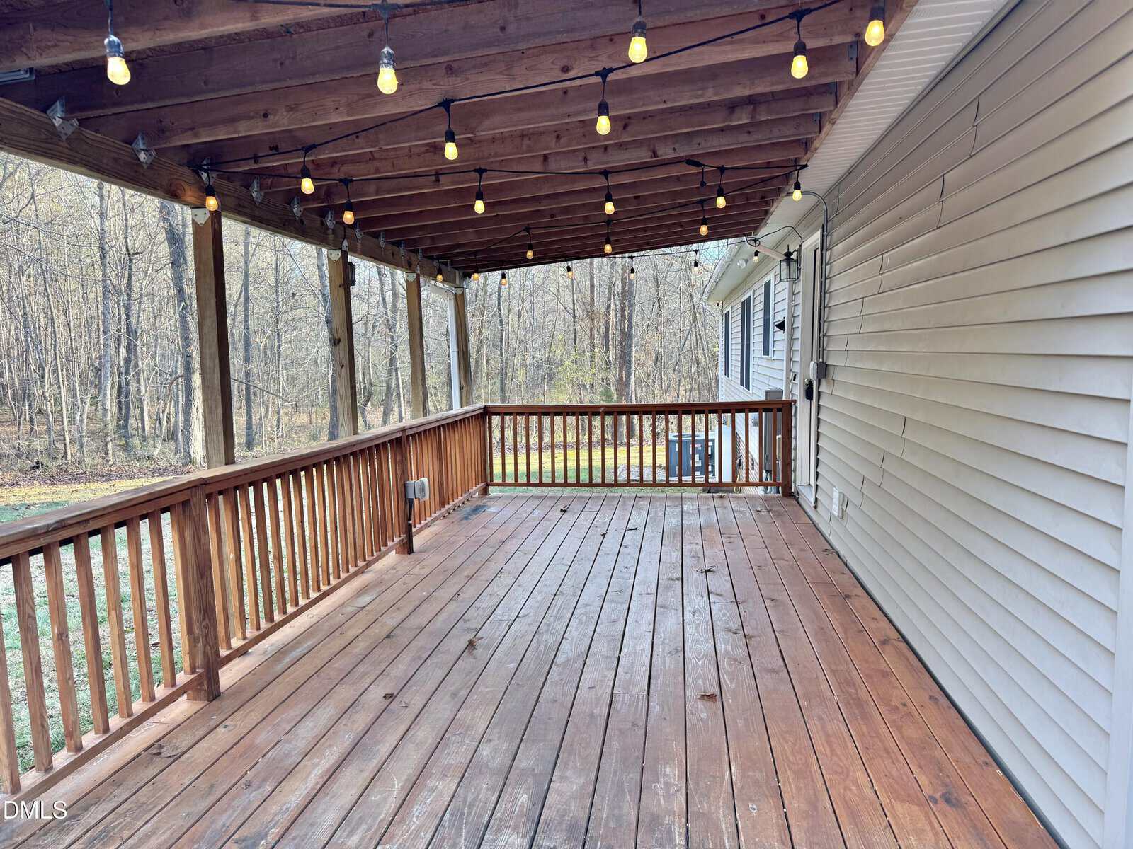 A covered wooden deck with string lights hanging from the ceiling, wooden railings, and a view of a wooded backyard. Siding of the house and part of a white door are visible on the right.