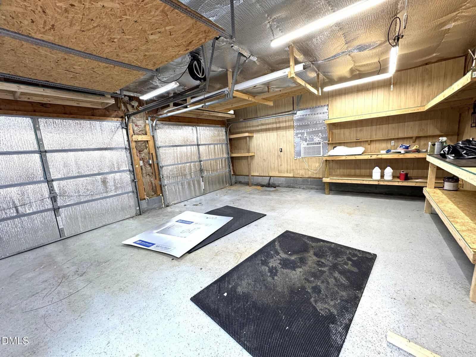A two-car garage with unfinished wooden walls, shelves, overhead lights, an air conditioning unit, black floor mats, and some items on the shelves. One garage door is partially open.