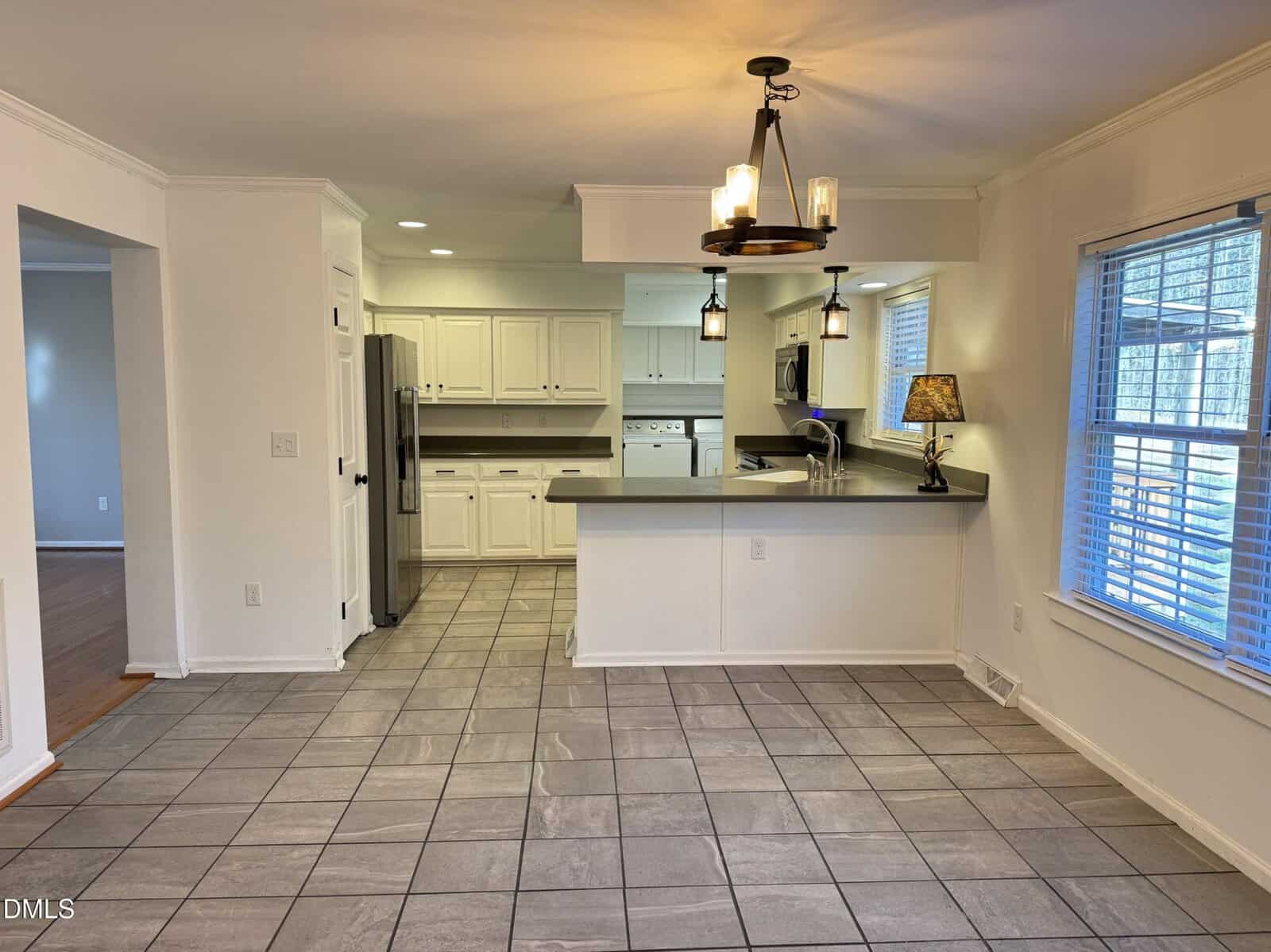 A kitchen with white cabinets, stainless steel appliances, gray countertops, and a pass-through bar. Tile flooring extends into a dining area with a hanging light fixture and a window with blinds.