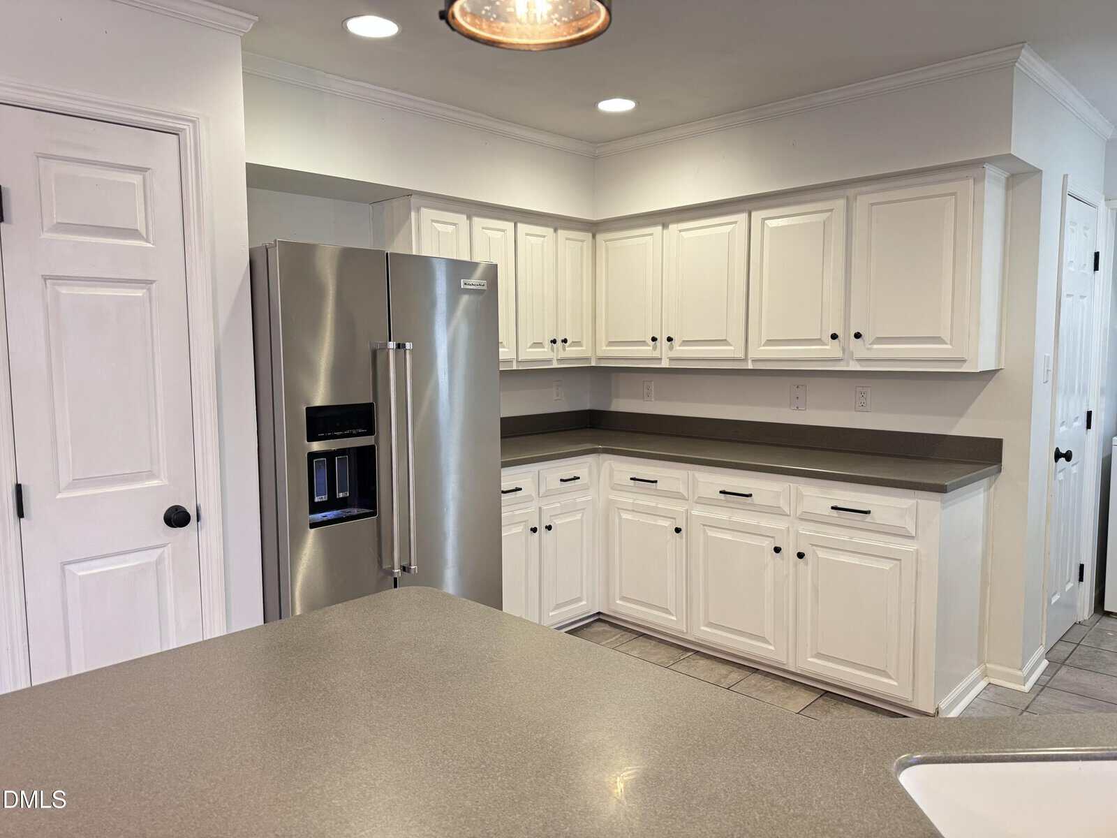 A modern kitchen with white cabinets, stainless steel refrigerator, gray countertops, tile flooring, and recessed lighting. The cabinets have black handles and there is a sink partially visible in the foreground.