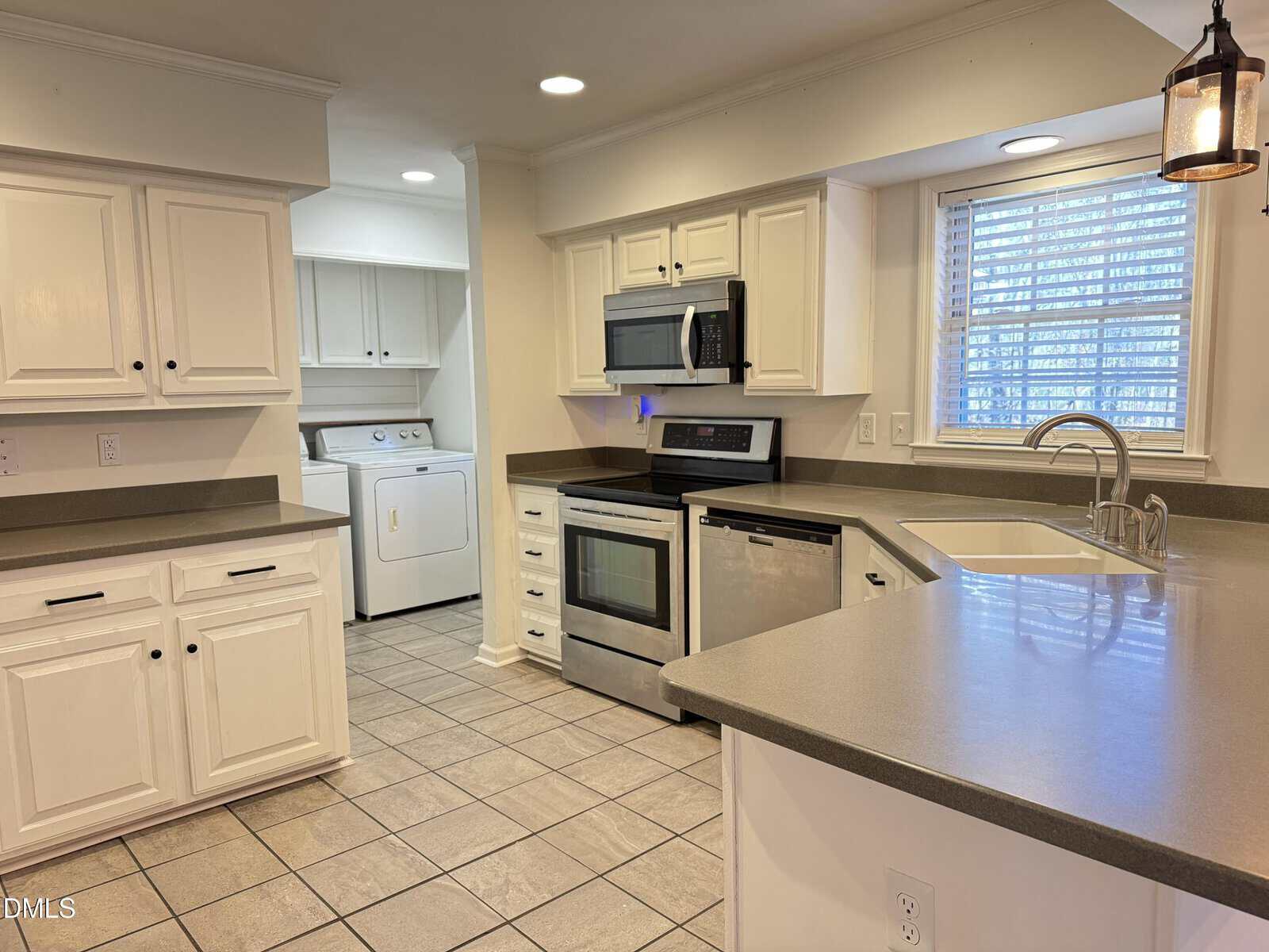 A kitchen with white cabinets, stainless steel appliances, and gray countertops. A built-in oven, microwave, dishwasher, and sink are visible, with a laundry area containing a washer and dryer in the adjacent room.