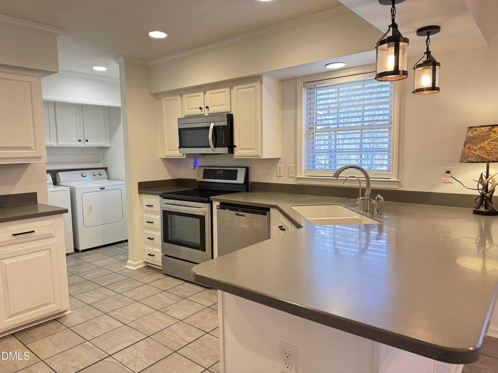 A modern kitchen with gray countertops, white cabinets, stainless steel appliances, a built-in microwave, a double sink, tile flooring, pendant lights, and a view into a laundry area with a washer and dryer.