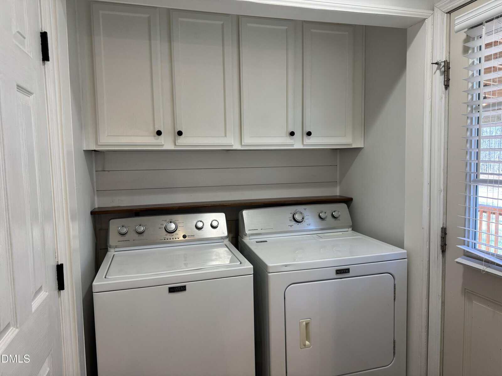 A laundry room with a white washing machine and dryer beneath white cabinets, next to a window with blinds.