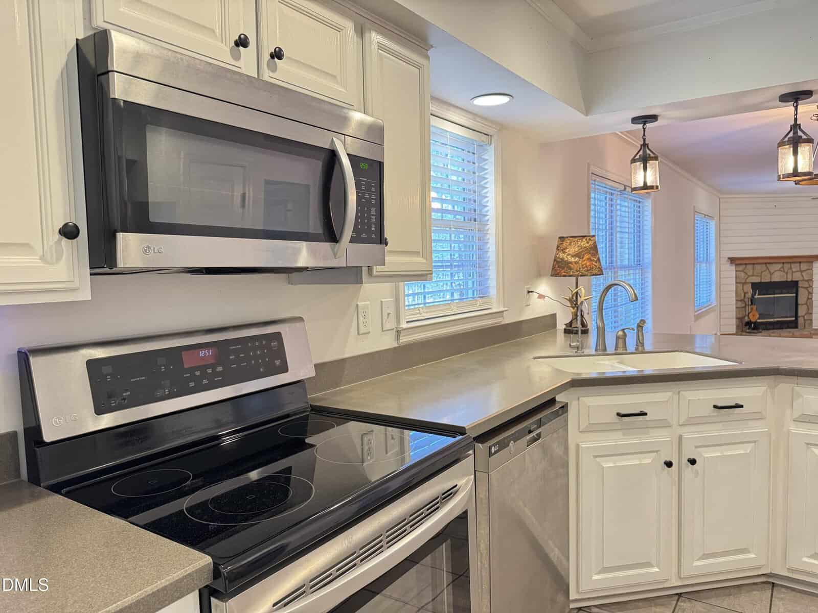 A kitchen with white cabinets, stainless steel appliances including a microwave and electric stove, a sink with a modern faucet, and a view of a living area with a fireplace and pendant lights.