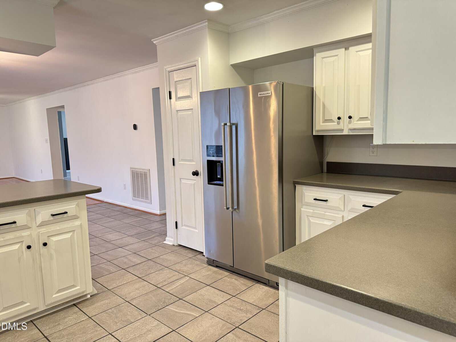 Modern kitchen with white cabinets, gray countertops, stainless steel refrigerator, and tile flooring. The view extends to a hallway with white walls and several doors visible in the background.