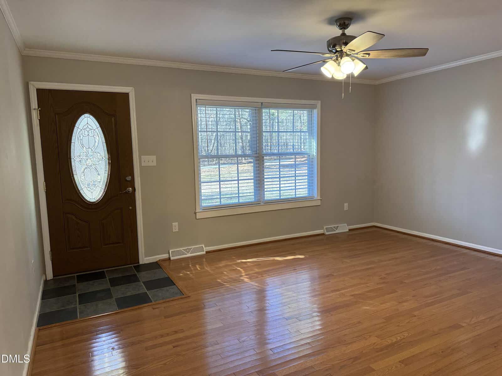 A vacant room with light brown hardwood floors, beige walls, a large window with blinds, a wooden front door with a decorative glass insert, and a ceiling fan with lights. There is a tiled entryway by the door.