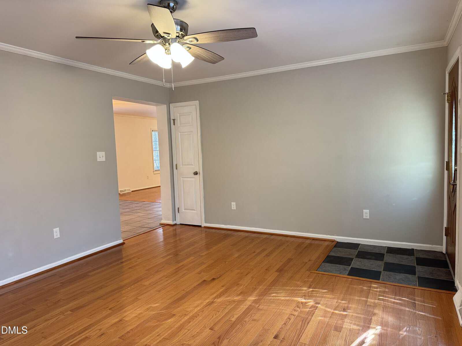 A room with light gray walls, a white ceiling, hardwood floor, ceiling fan with lights, a small tiled entryway, and a doorway leading to another room with tile flooring.