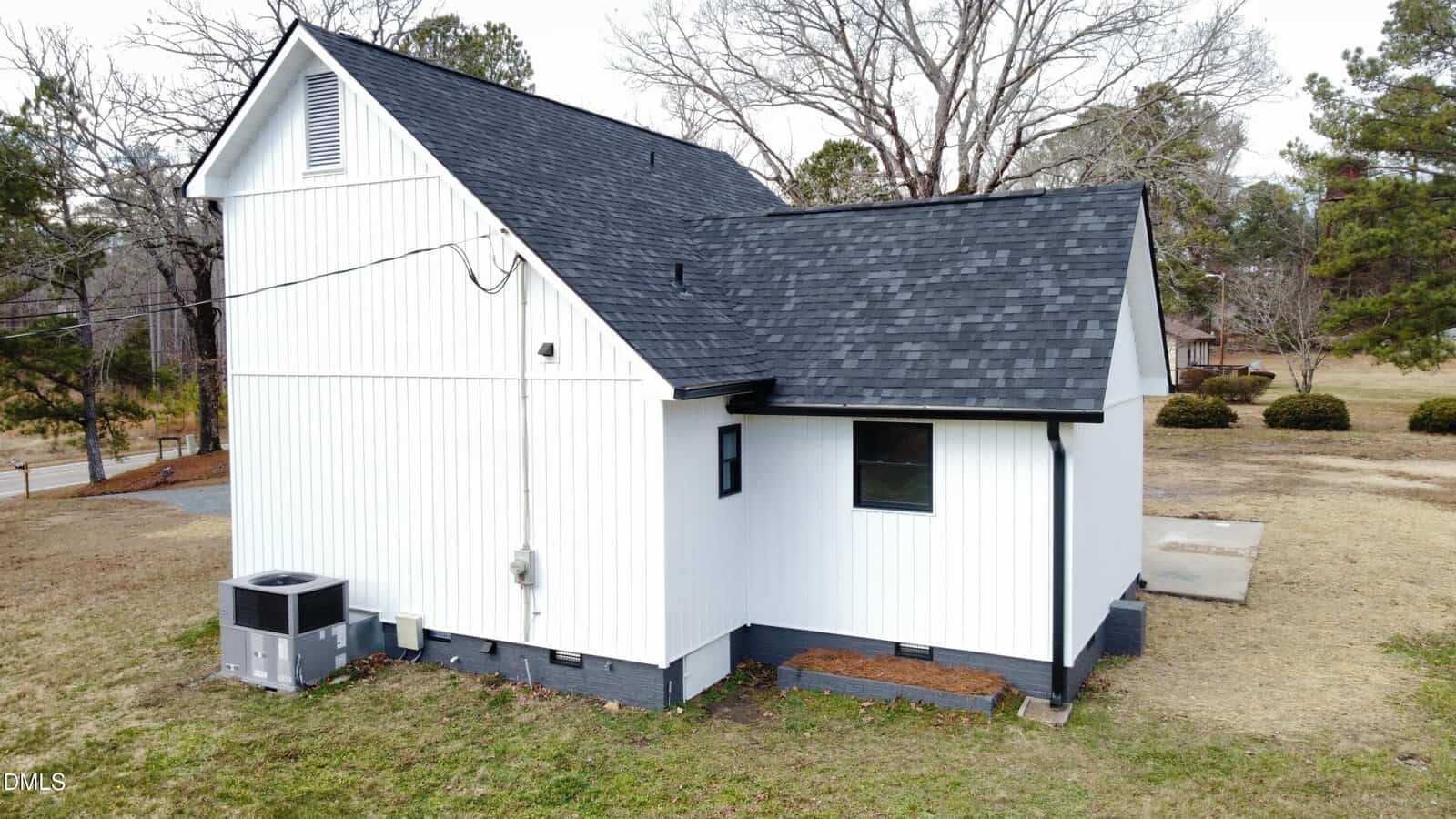 A white two-story house with black shingles, black trim, and a gray foundation sits on a grassy lot. An HVAC unit is beside the house, and leafless trees are visible in the background.