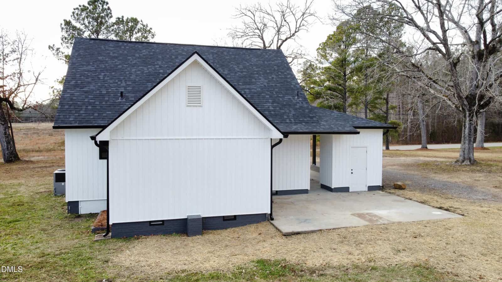 A small white house with black trim and a dark shingled roof, featuring a covered carport and an adjacent storage room, sits on a grassy lot with trees and a dirt driveway in the background.