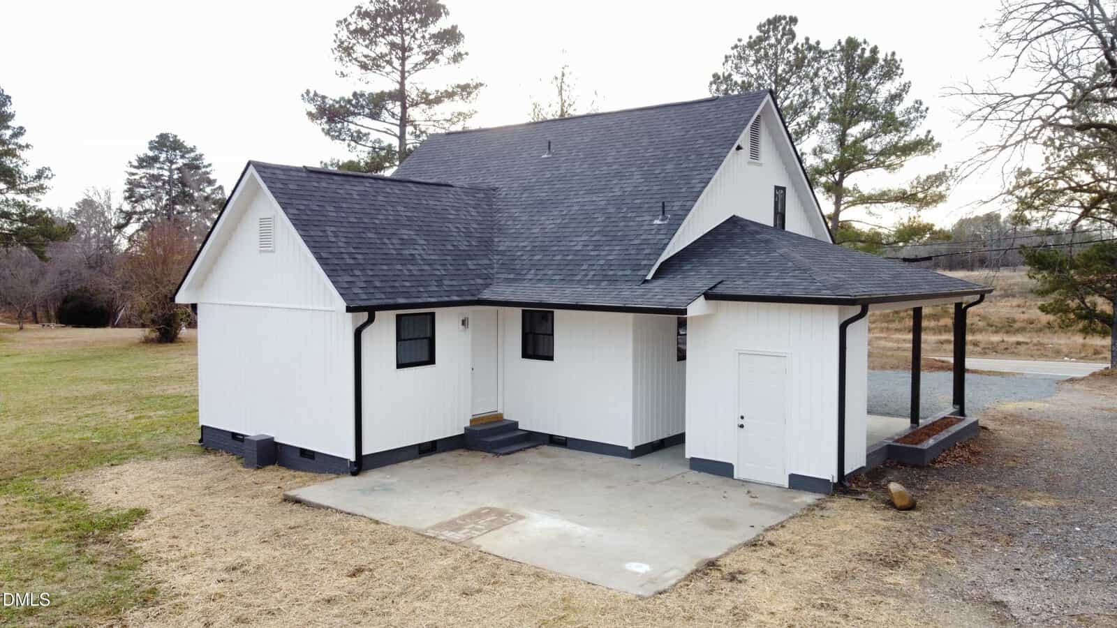 A white two-story house with black trim and a dark gray roof sits on a plot with patchy grass. There is a small covered porch, a concrete slab, and several trees in the background.