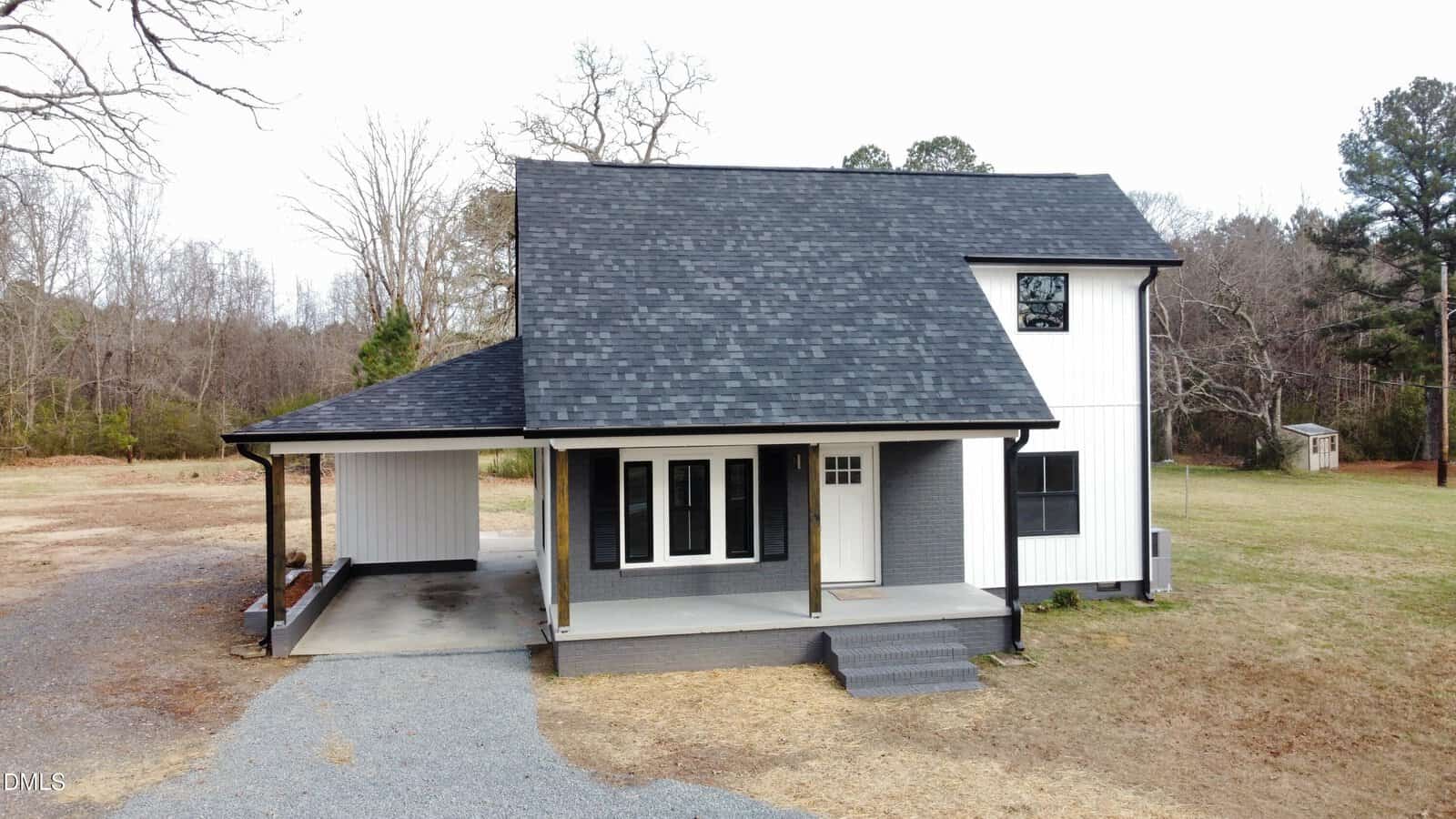 A modern two-story house with black and white exterior, black roof, covered carport, front porch with columns, and surrounded by grass, gravel driveway, and trees in the background.