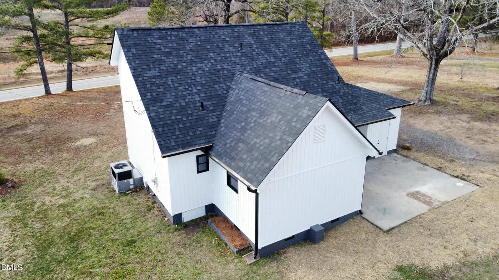 A white house with a dark shingled roof sits on a grassy lot bordered by trees and a road. There is an air conditioning unit, a small window, and a concrete pad next to the house.