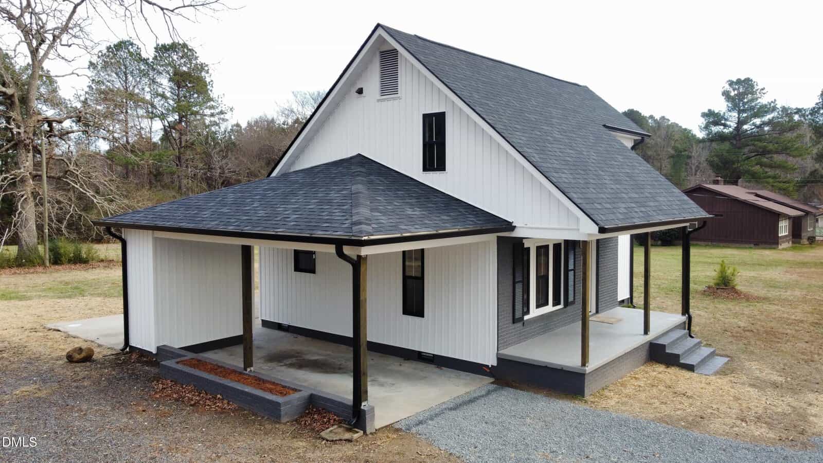 A modern white house with black trim, a covered carport, front porch, and dark shingled roof. The house sits on a grassy lot with trees and another building visible in the background.