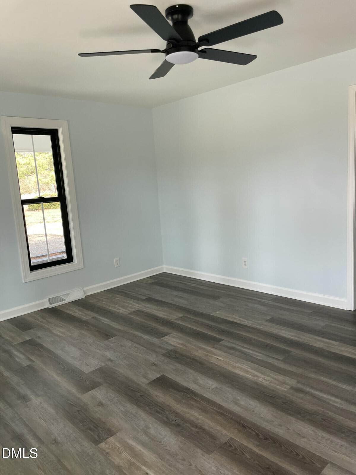 Empty room with light blue walls, a black ceiling fan, gray wood-style flooring, a single window with black trim, and white baseboards. Sunlight enters from the window, creating a bright atmosphere.