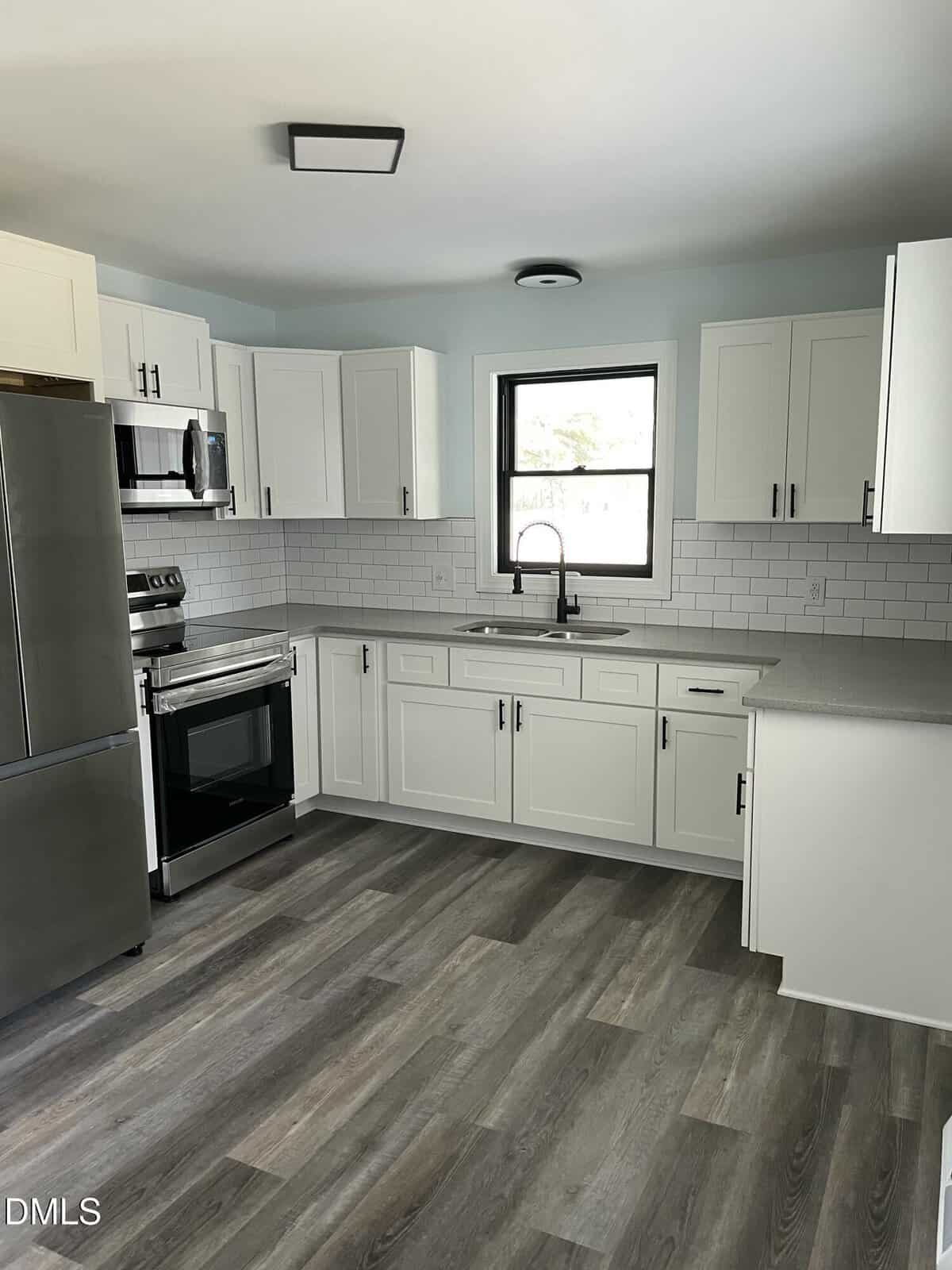 Modern kitchen with white cabinets, stainless steel appliances, subway tile backsplash, gray countertops, a single window above the sink, and gray wood-look flooring.