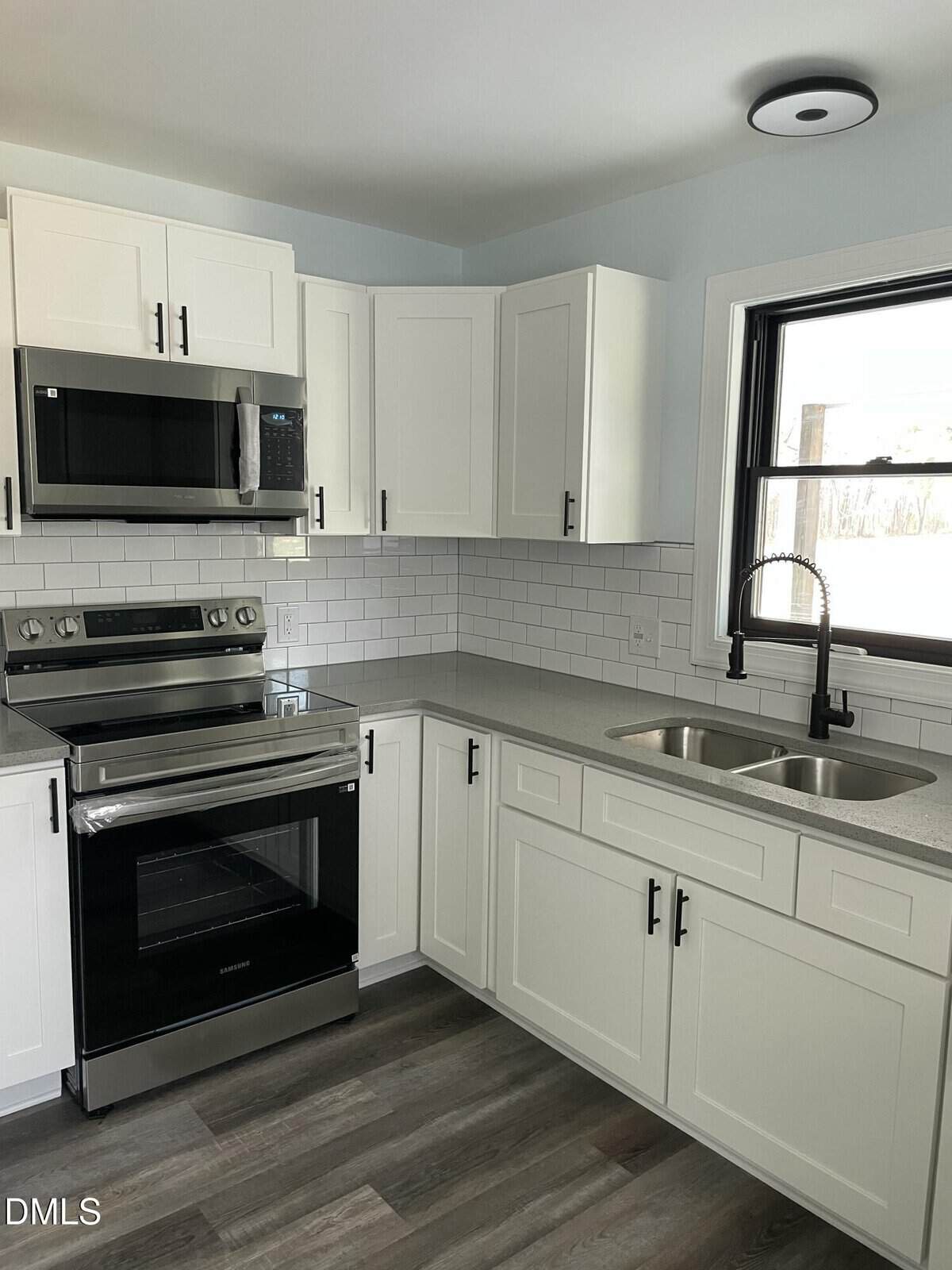 Modern kitchen with white cabinets, stainless steel appliances, subway tile backsplash, gray countertops, double sink with a tall faucet, and a window above the sink. The floor is wood-style laminate.