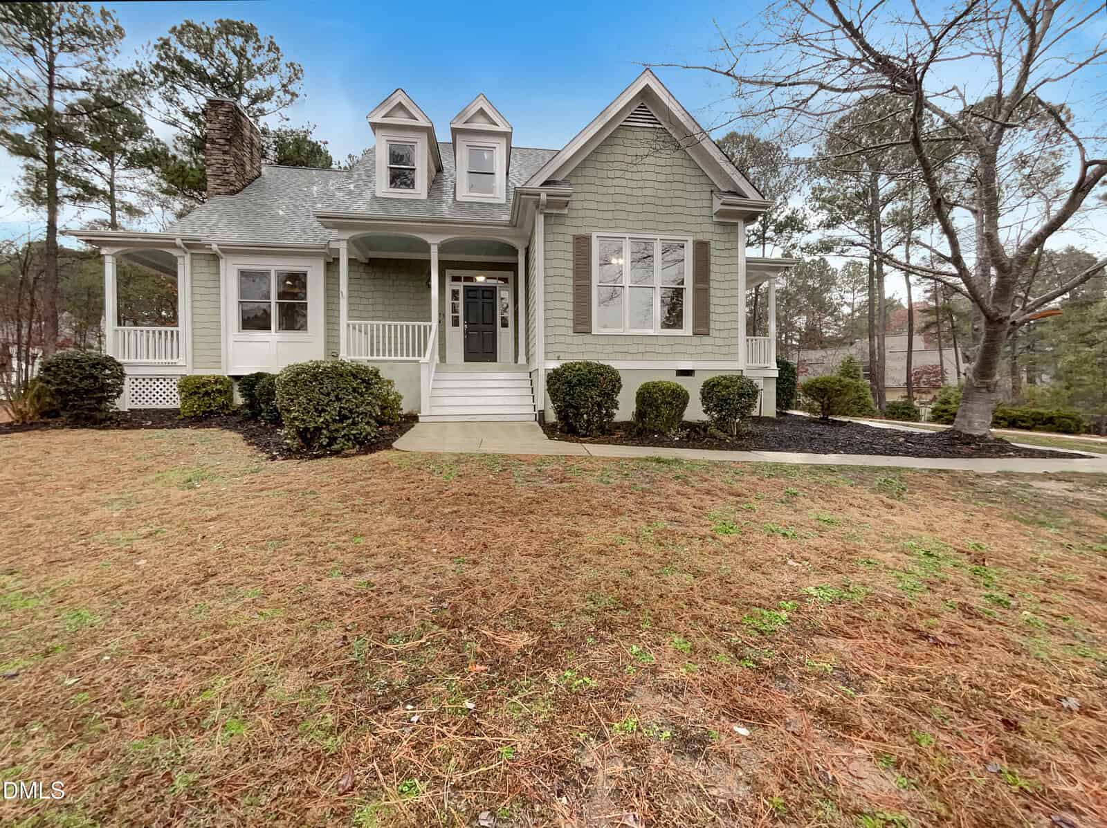 A light green two-story house with white trim, a covered front porch, dormer windows, and a sloped yard with shrubs and pine straw, surrounded by trees under a blue sky.