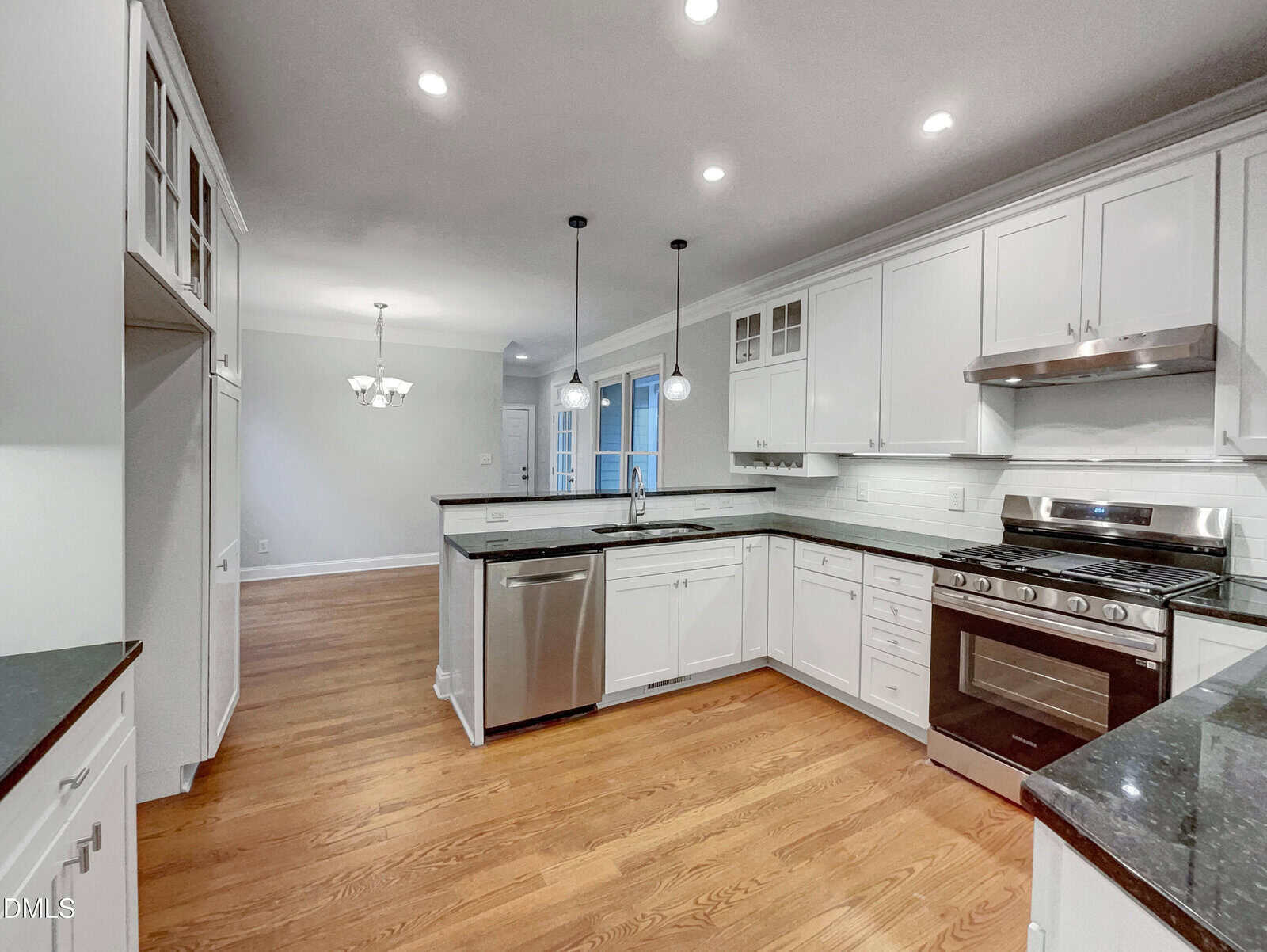 Modern kitchen with white cabinets, stainless steel appliances, granite countertops, wood floors, and pendant lighting. Open layout leads to a dining area with a chandelier in the background.