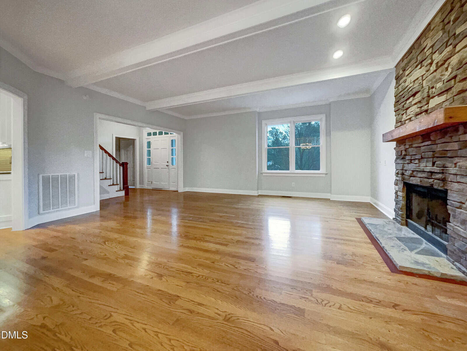 Spacious, empty living room with light hardwood floors, white walls, recessed ceiling lights, a large window, a stone fireplace, and a staircase near the entryway.