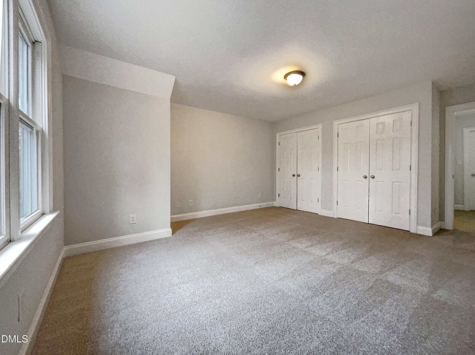 Spacious empty room with light gray walls, beige carpet, a ceiling light fixture, two closets with double doors, and large windows letting in natural light.