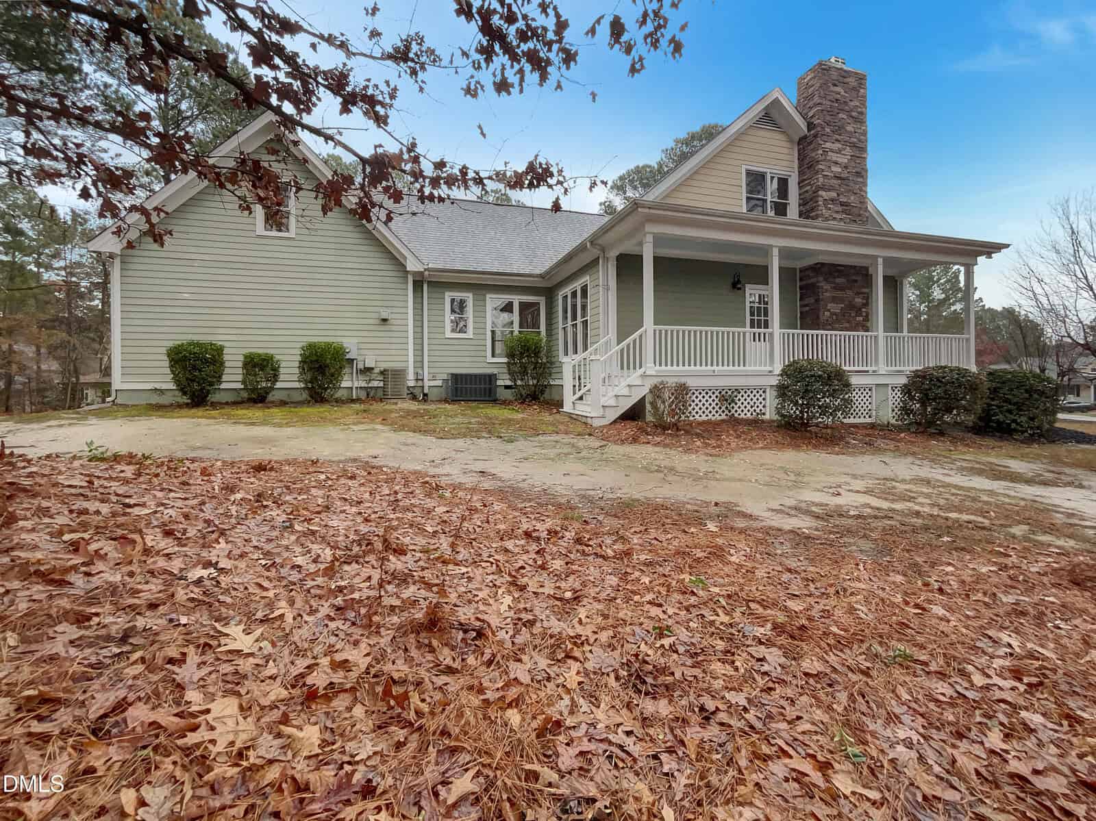 A green house with a large front porch, stone chimney, and white railings, surrounded by trees and fallen leaves on the ground under a blue sky.