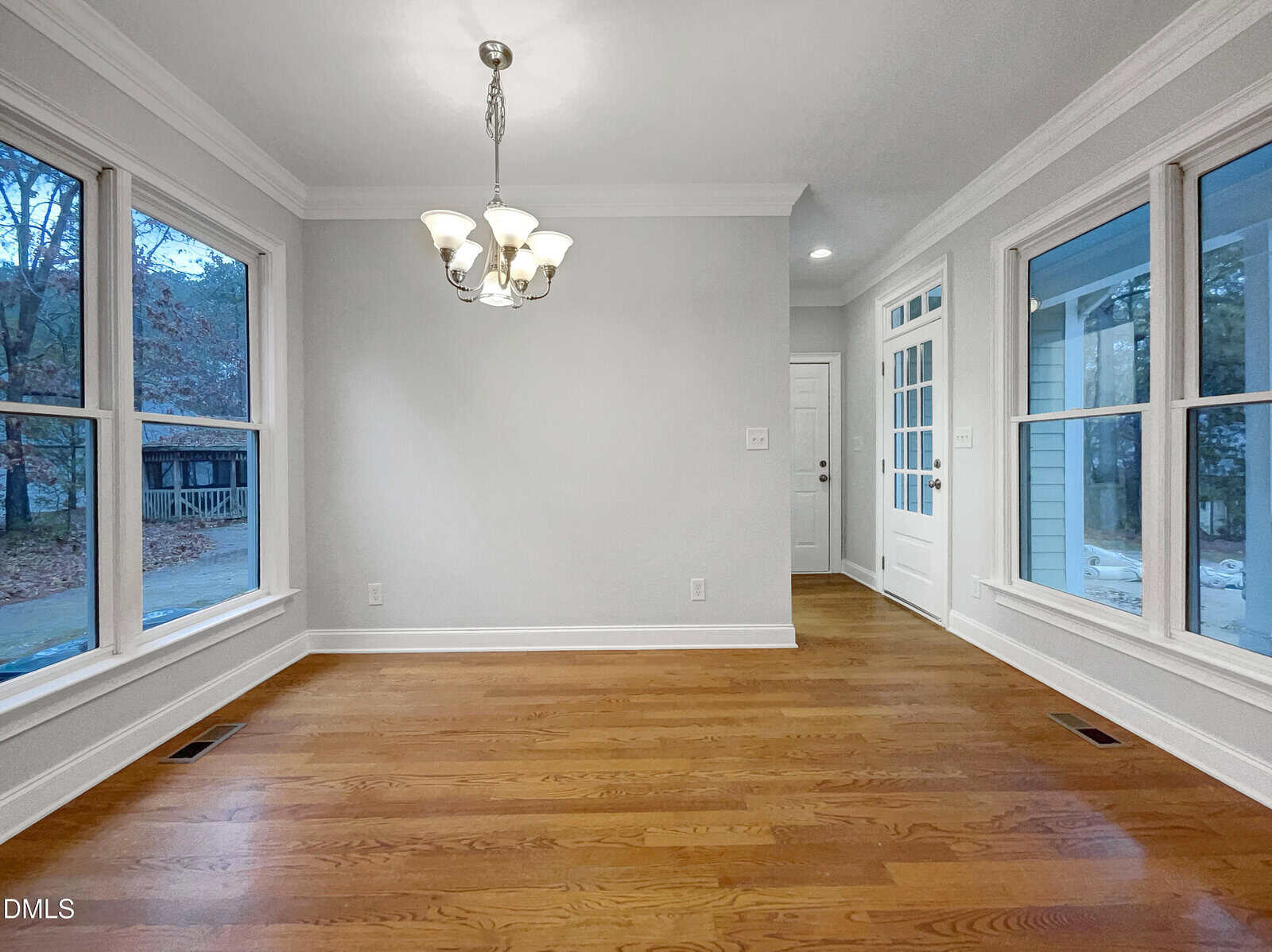A bright, empty dining room with hardwood floors, large windows on two walls, a white door, and a modern chandelier hanging from the ceiling. Natural light fills the space.