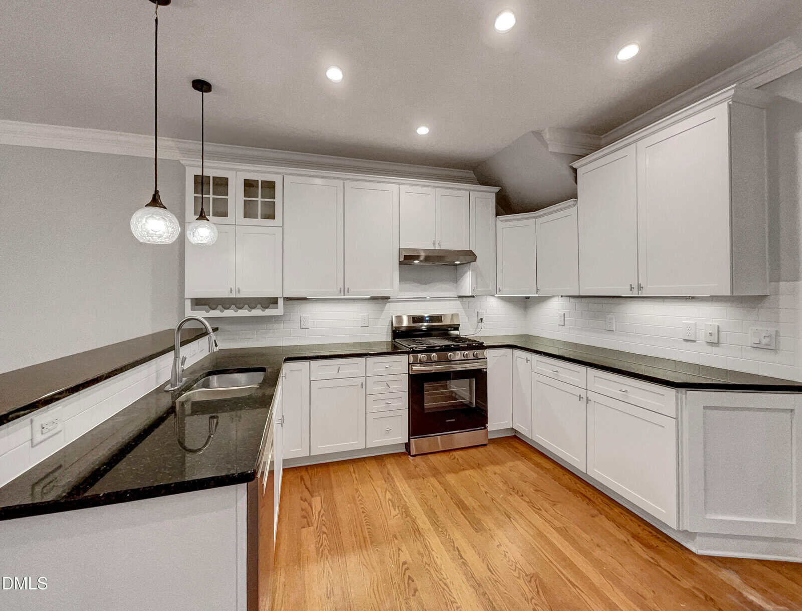 Modern kitchen with white cabinets, black countertops, stainless steel appliances, a gas stove, wood flooring, and pendant lights over a peninsula with a sink. Brightly lit with recessed ceiling lights.