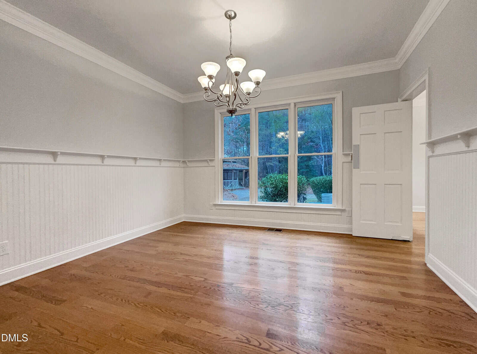 A bright, empty dining room with hardwood floors, white wainscoting, gray walls, a large window, a chandelier, and a white door partially open to the right.