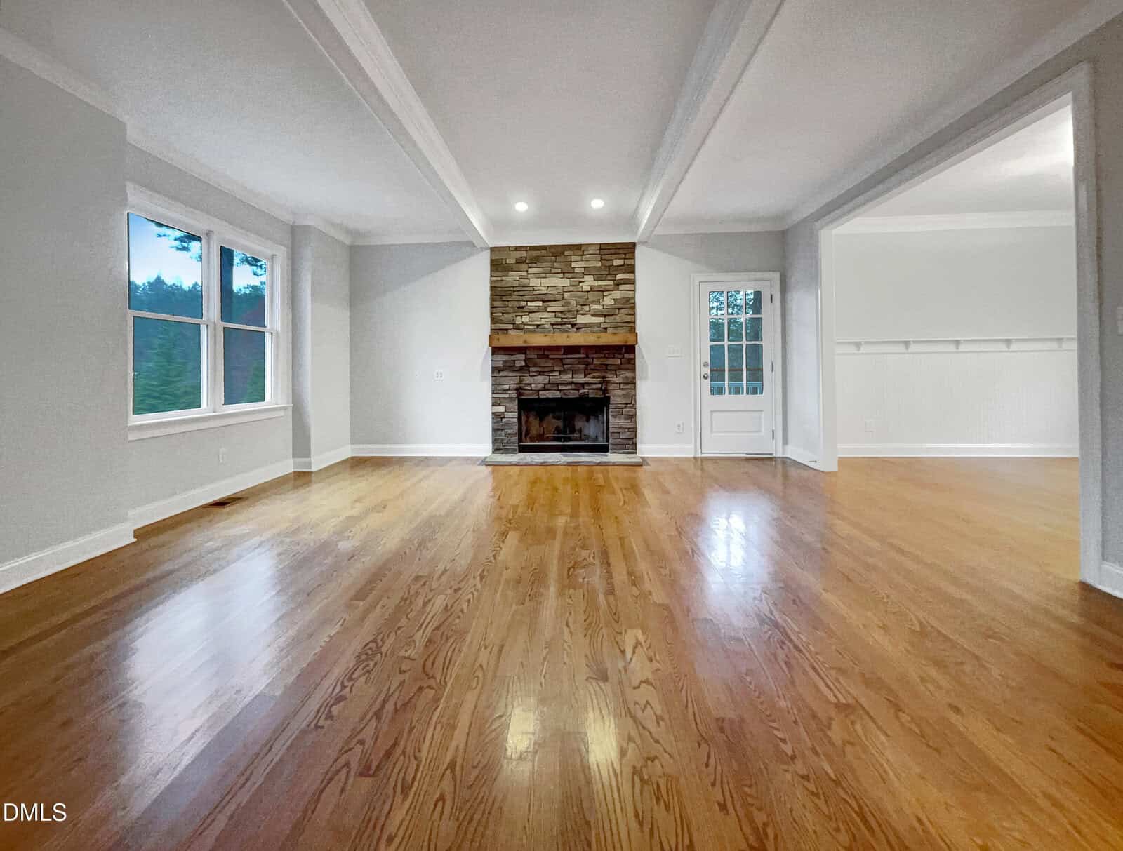 Spacious living room with polished wood floors, white walls, large windows, a stone fireplace with a mantel, ceiling beams, and an open doorway leading to another room with similar flooring.