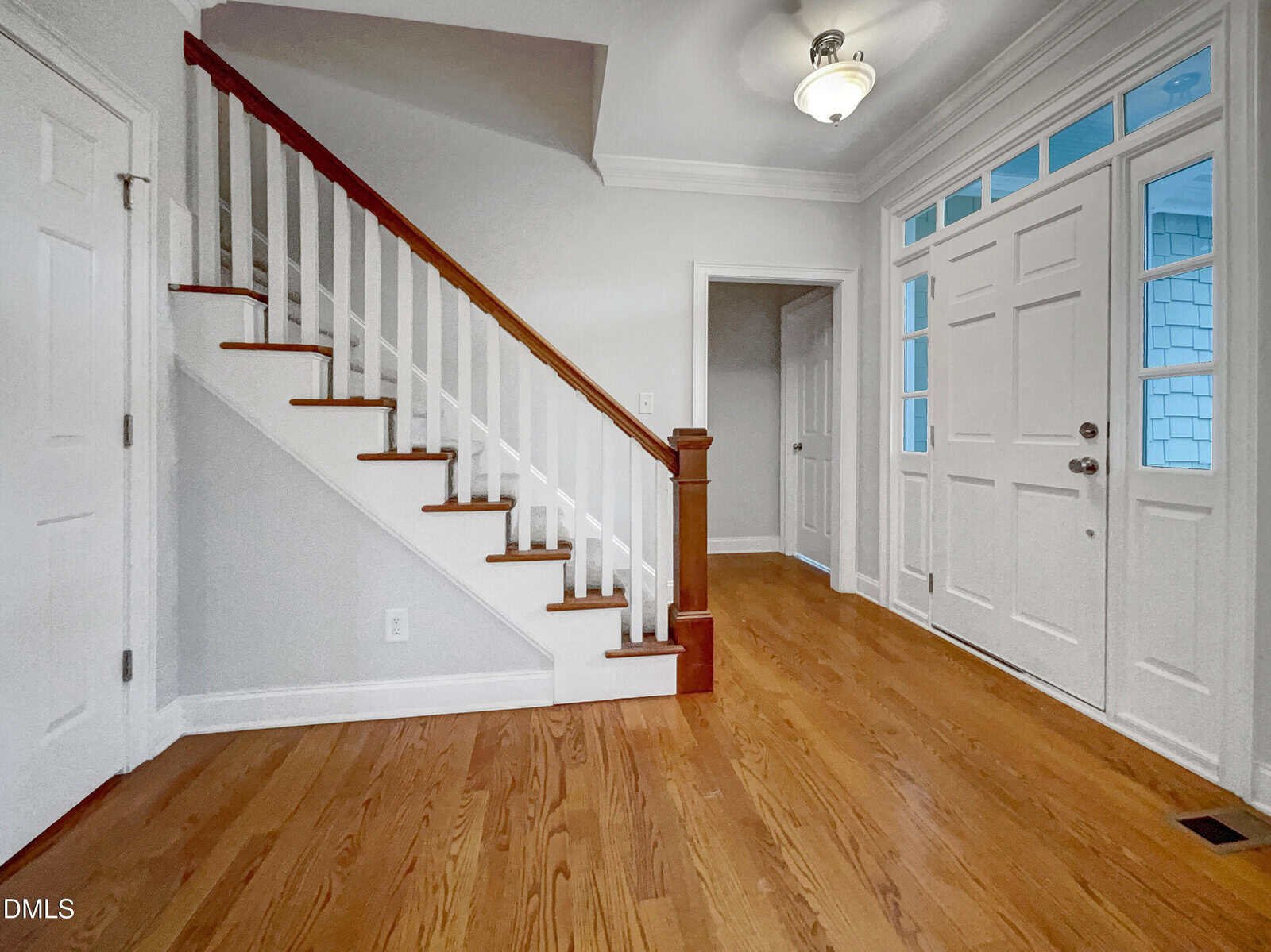 A bright entryway with hardwood floors, white walls, a wooden staircase with white railings, and a white front door with windows above and beside it. A ceiling light fixture hangs overhead.