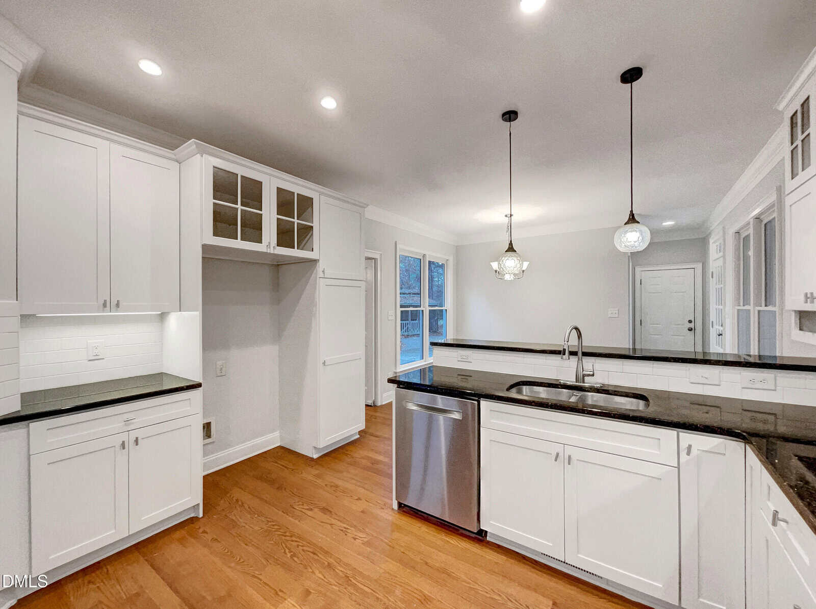 Bright modern kitchen with white cabinets, black countertops, stainless steel appliances, pendant lights over a large island, and wood flooring. Large windows and recessed lighting create an airy, spacious feel.