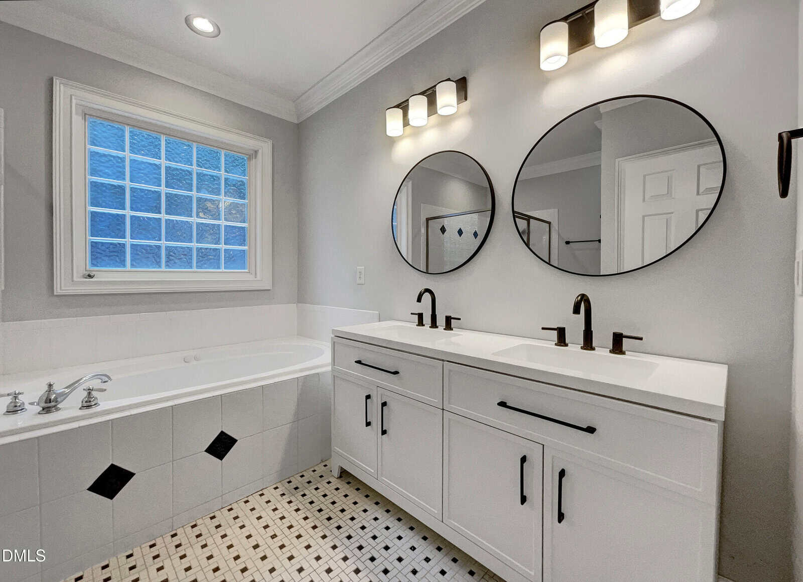 Modern bathroom with a white double vanity, two round mirrors, black fixtures, a built-in bathtub with silver faucets, and a patterned tile floor. A window above the tub lets in natural light.