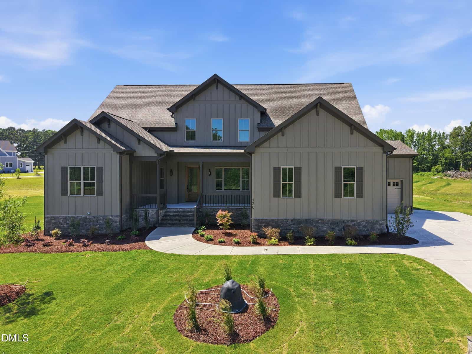 A modern, gray two-story house with a gabled roof, stone accents, a covered front porch, and a neatly landscaped lawn under a bright blue sky. A curved driveway and a small garden with shrubs are visible in front.