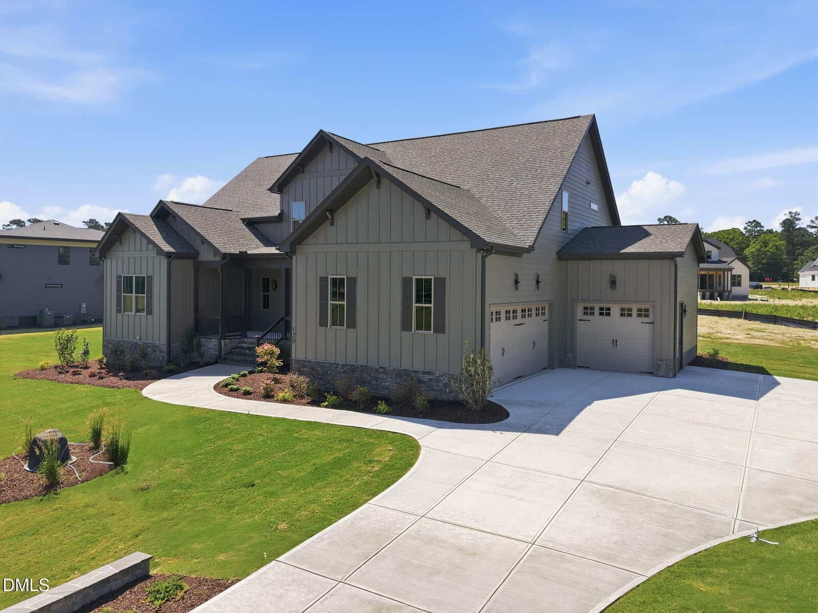 A modern, gray house with gabled roofs, stone accents, and a three-car garage, sits on a neatly landscaped lawn with a curved driveway under a clear blue sky.
