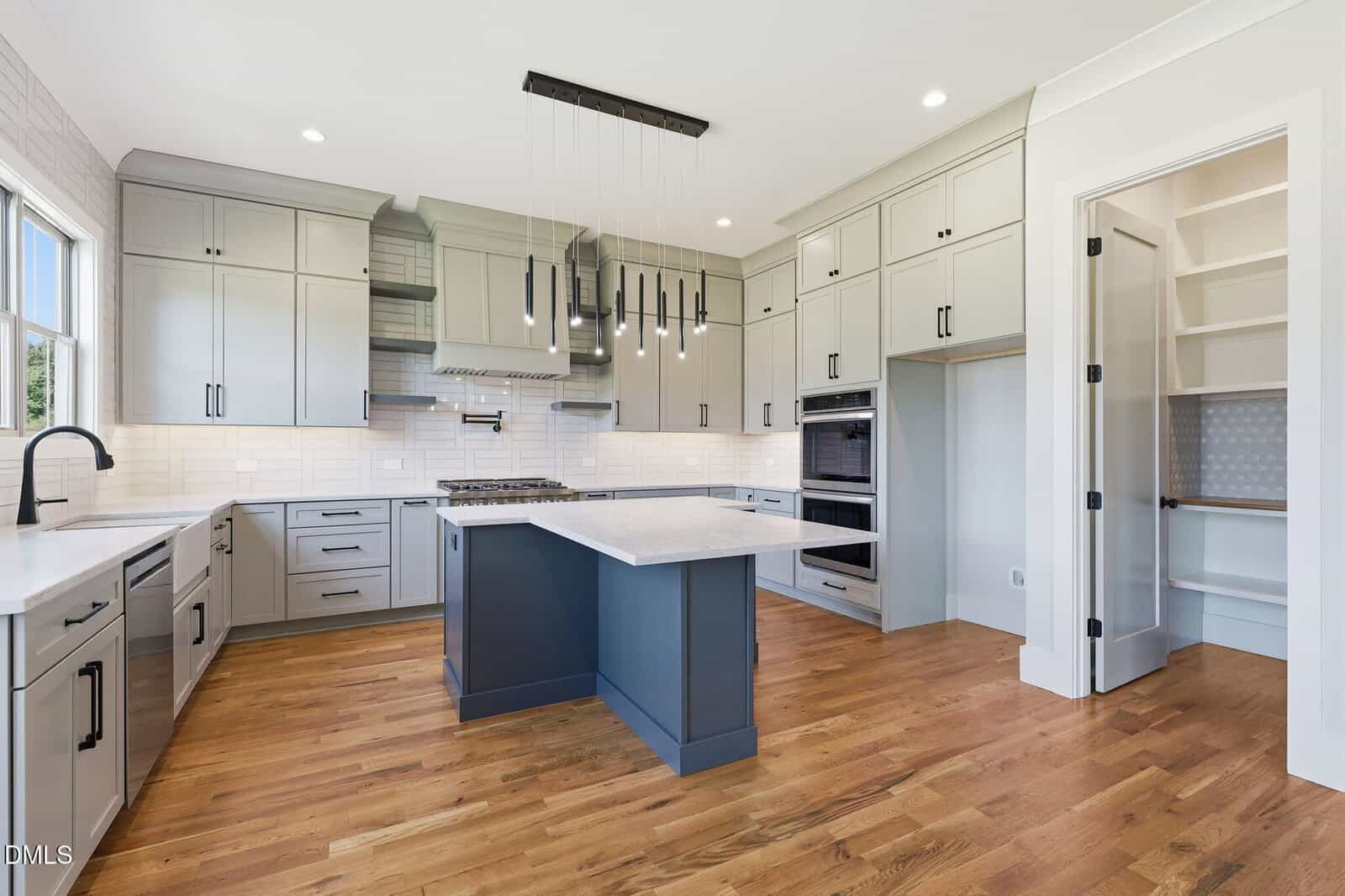 Modern kitchen with light gray cabinets, a blue island, stainless steel appliances, pendant lights, wood floors, large windows, and a walk-in pantry with built-in shelves.