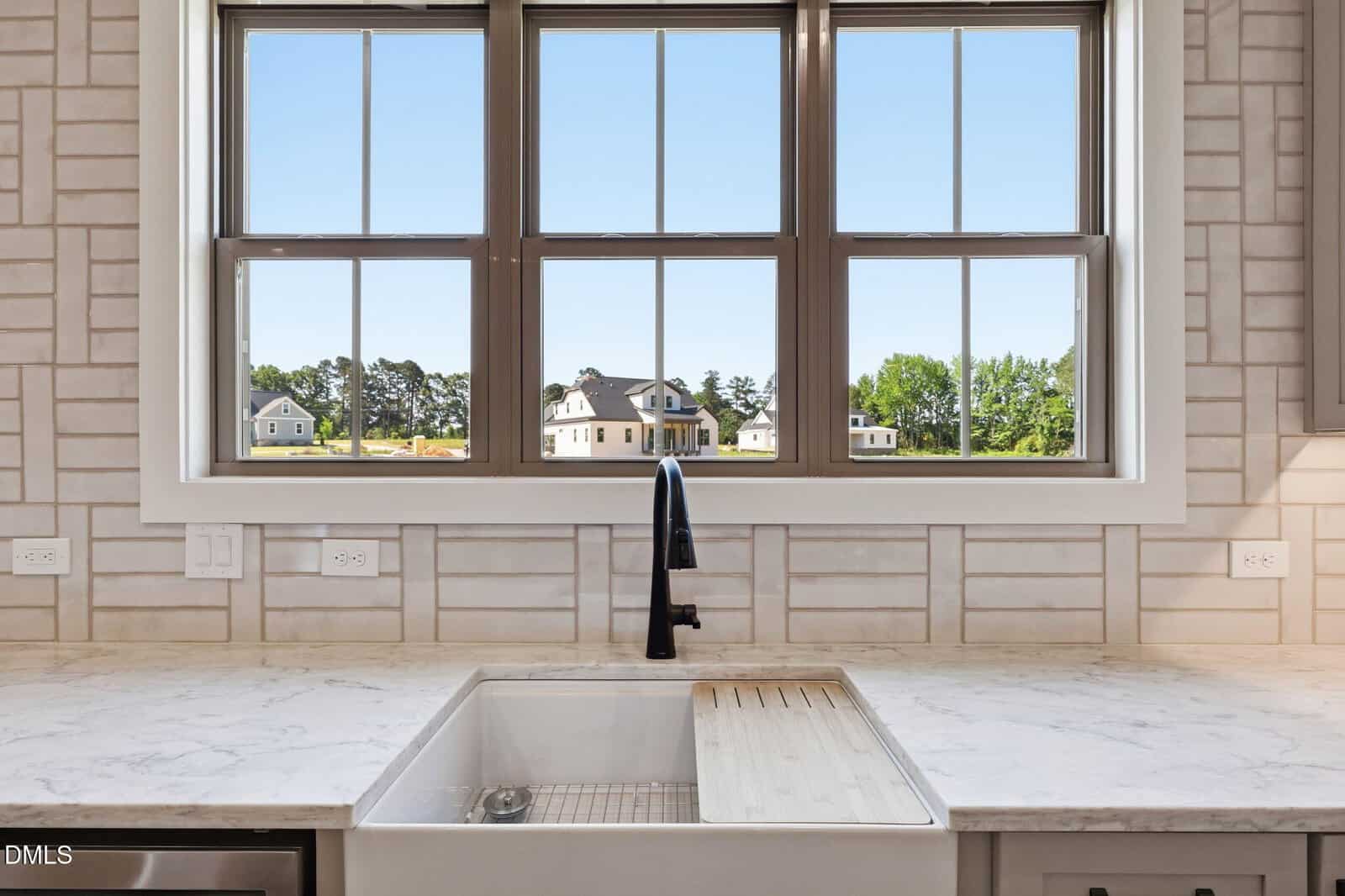 A white farmhouse sink with a black faucet sits below a large window, offering a clear view of houses, green lawns, and trees outside on a sunny day. Marble countertops and white tiled walls frame the kitchen.