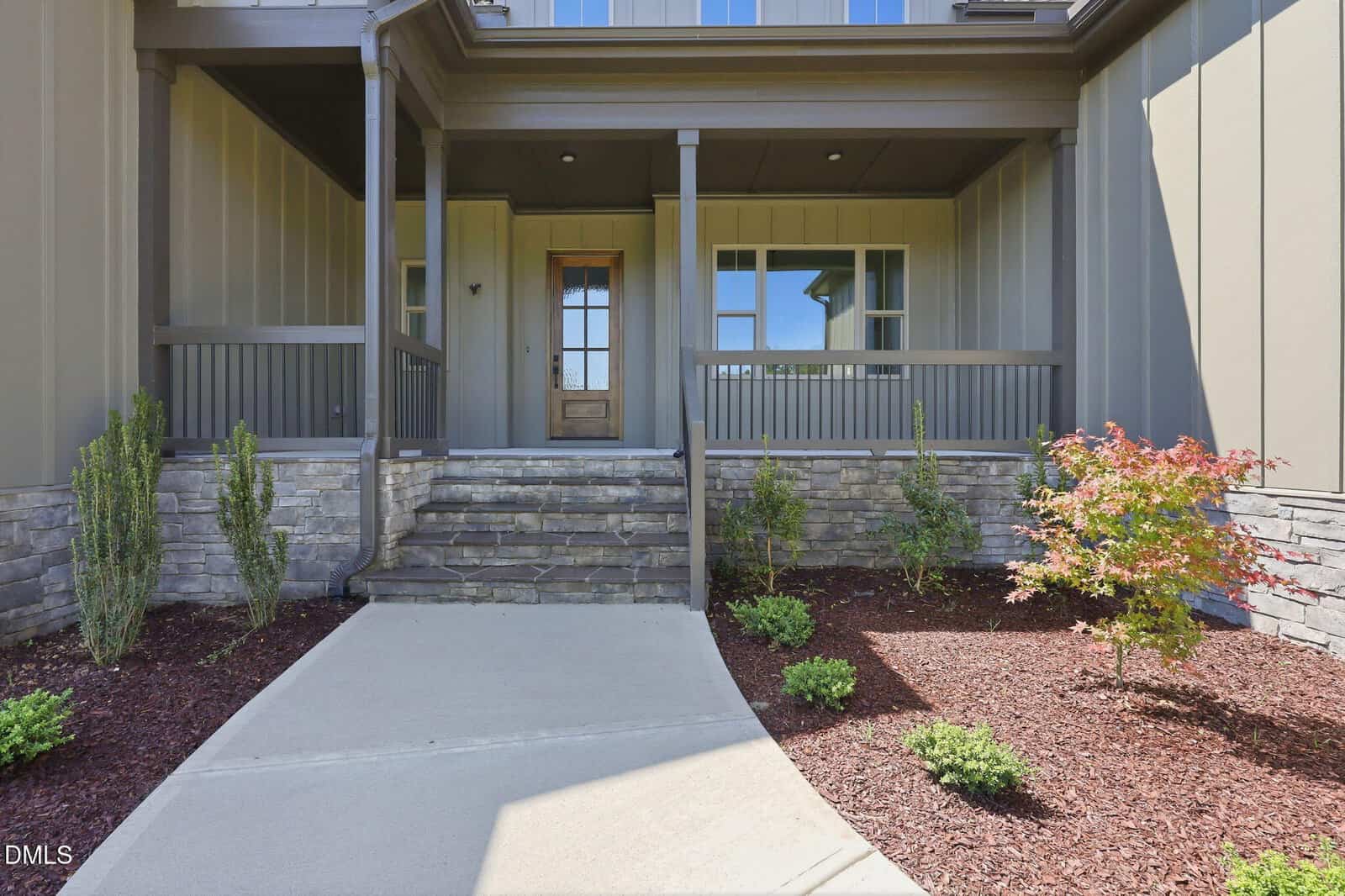 A modern house entrance with a wooden front door, stone steps, covered porch, and landscaped yard featuring small shrubs and a small tree beside a concrete walkway.