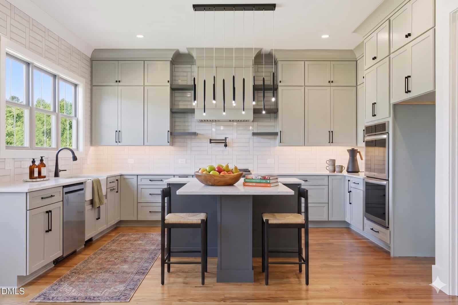 Modern kitchen with light gray cabinets, stainless steel appliances, a black island with two stools, a bowl of fruit, wood floors, a rug, and large windows letting in natural light.