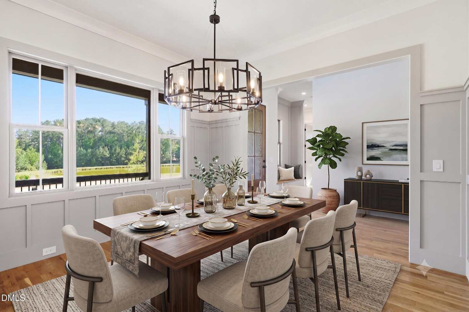 A bright dining room with a large wooden table set for eight, beige upholstered chairs, a modern chandelier, and large windows overlooking greenery. The room opens to a living area with a plant and artwork.