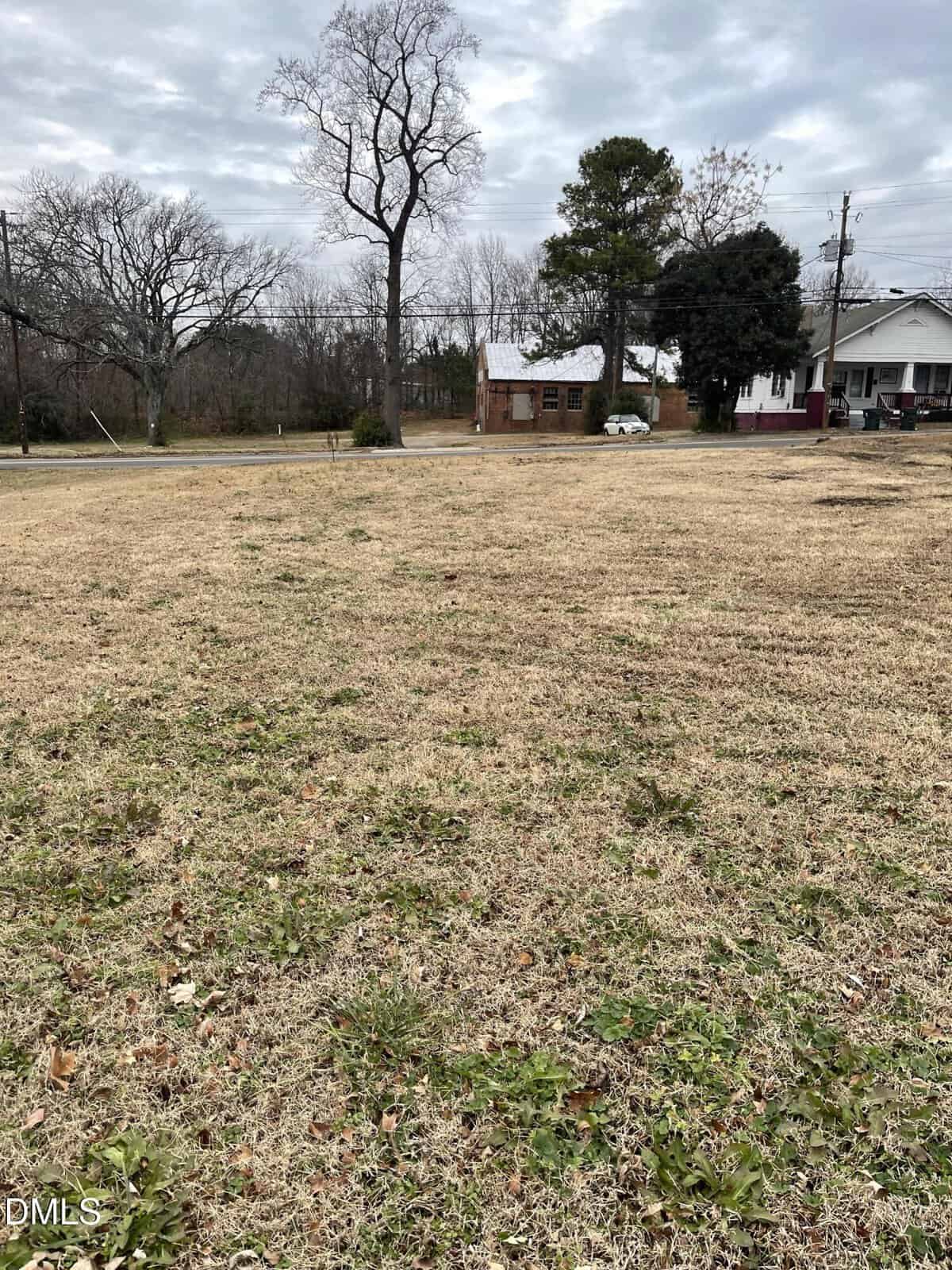 A grassy vacant lot with sparse green and brown patches, bordered by a road on 507 Hillsboro Street in Oxford. Leafless trees and two houses are visible in the background under a cloudy sky.