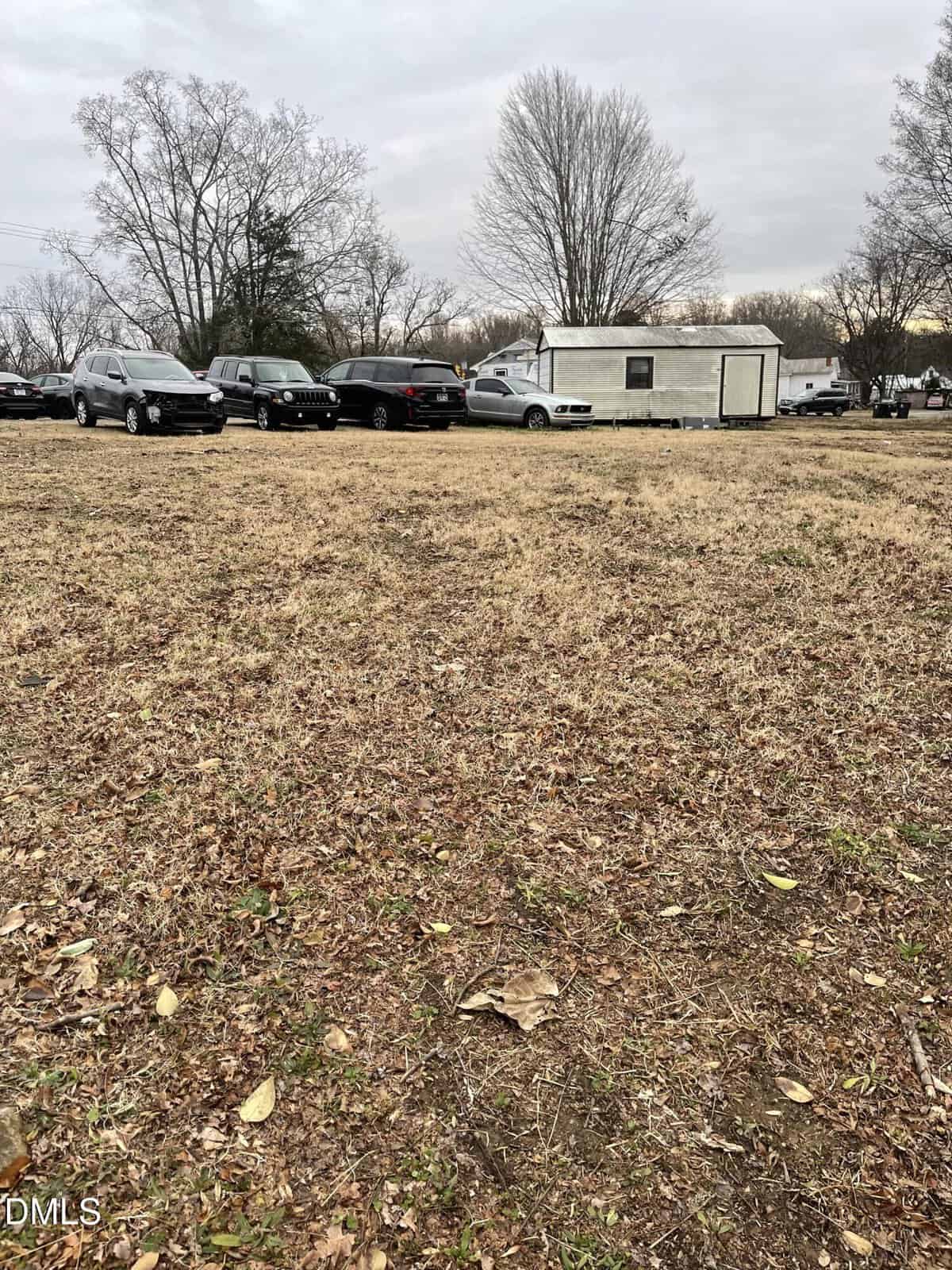 A mostly bare, grassy lot with scattered leaves in the foreground. Several parked cars and a white trailer or mobile building are visible in the background, along with leafless trees under a cloudy sky.