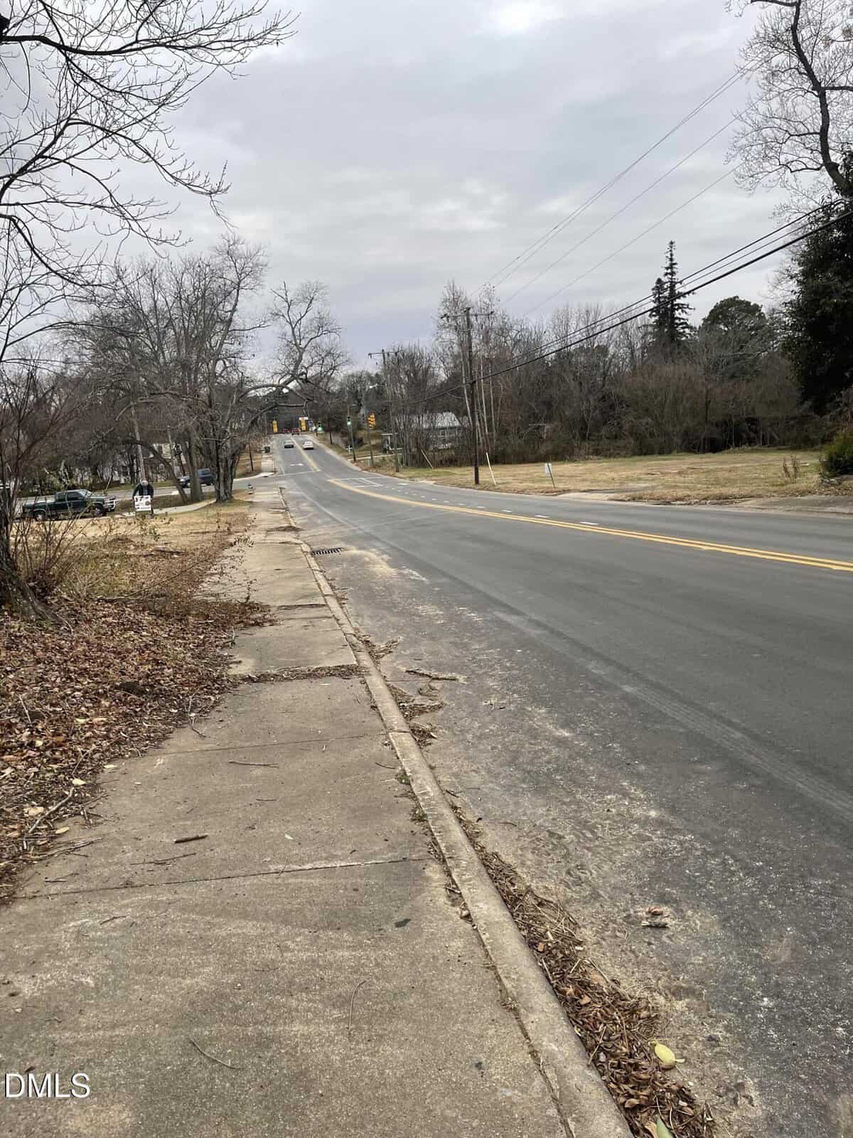 A two-lane road runs through a quiet, mostly empty neighborhood. The sidewalk is cracked, with bare trees and dry grass lining both sides. Overhead power lines are visible under a cloudy sky.
