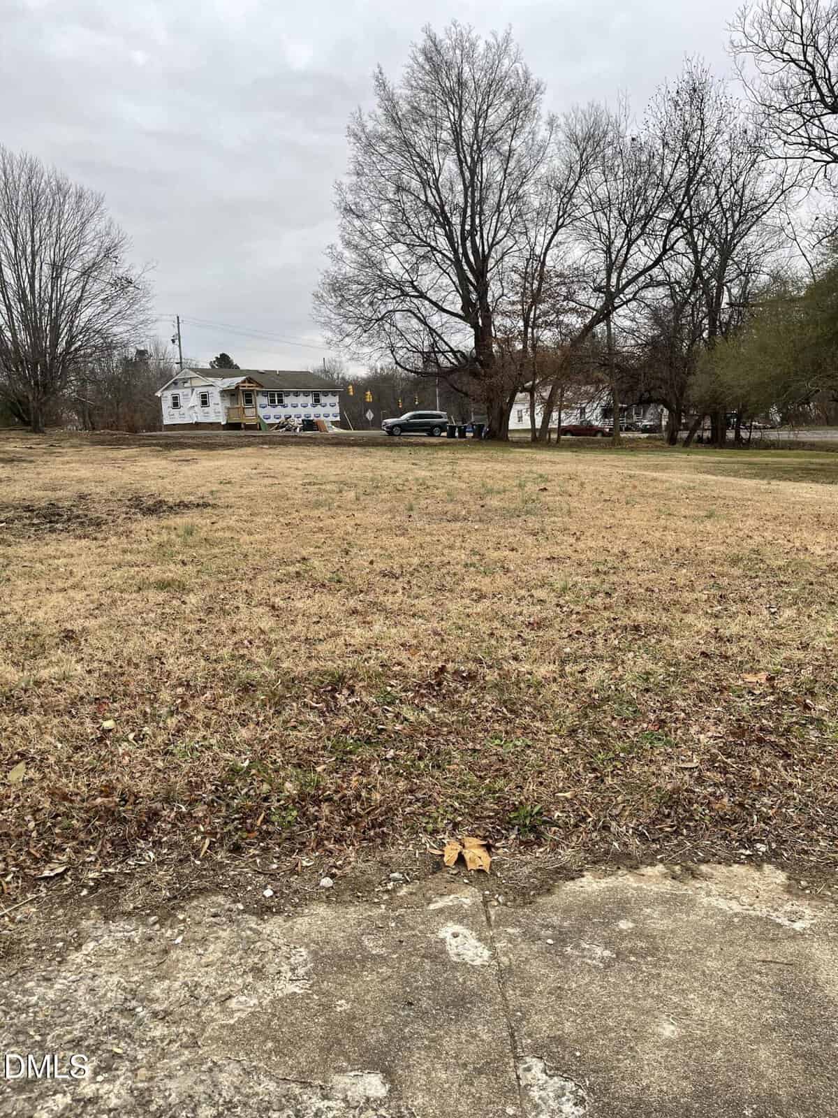 A vacant lot with dry grass and a few scattered leaves in the foreground. Bare trees line the back, and houses and a parked car are visible across the street under a cloudy sky in Oxford, near 513 Hillsboro Street.